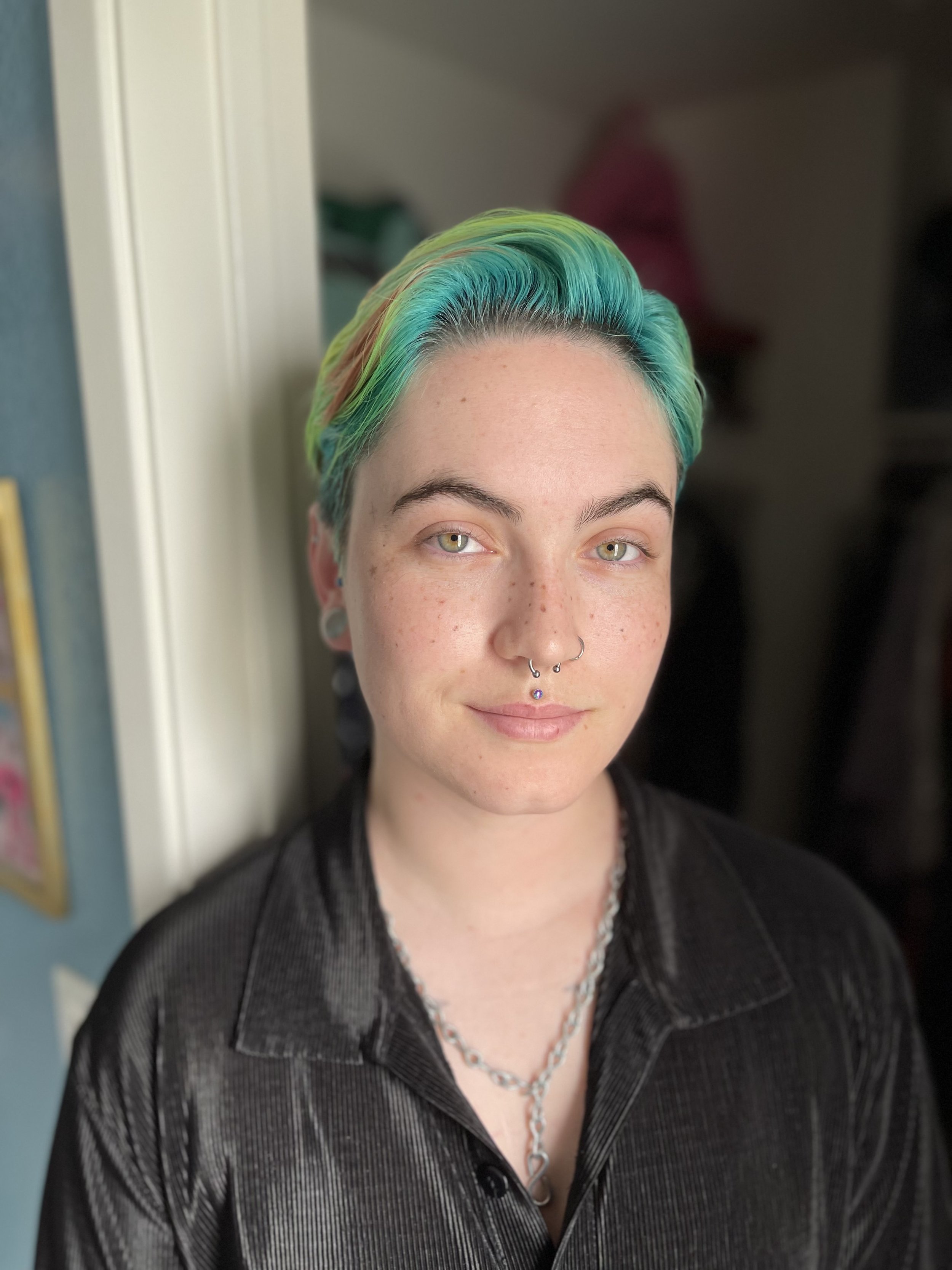 A person with short rainbow-colored hair, light skin, and multiple facial piercings, wearing a black jacket and silver jewelry, standing indoors near a doorway.