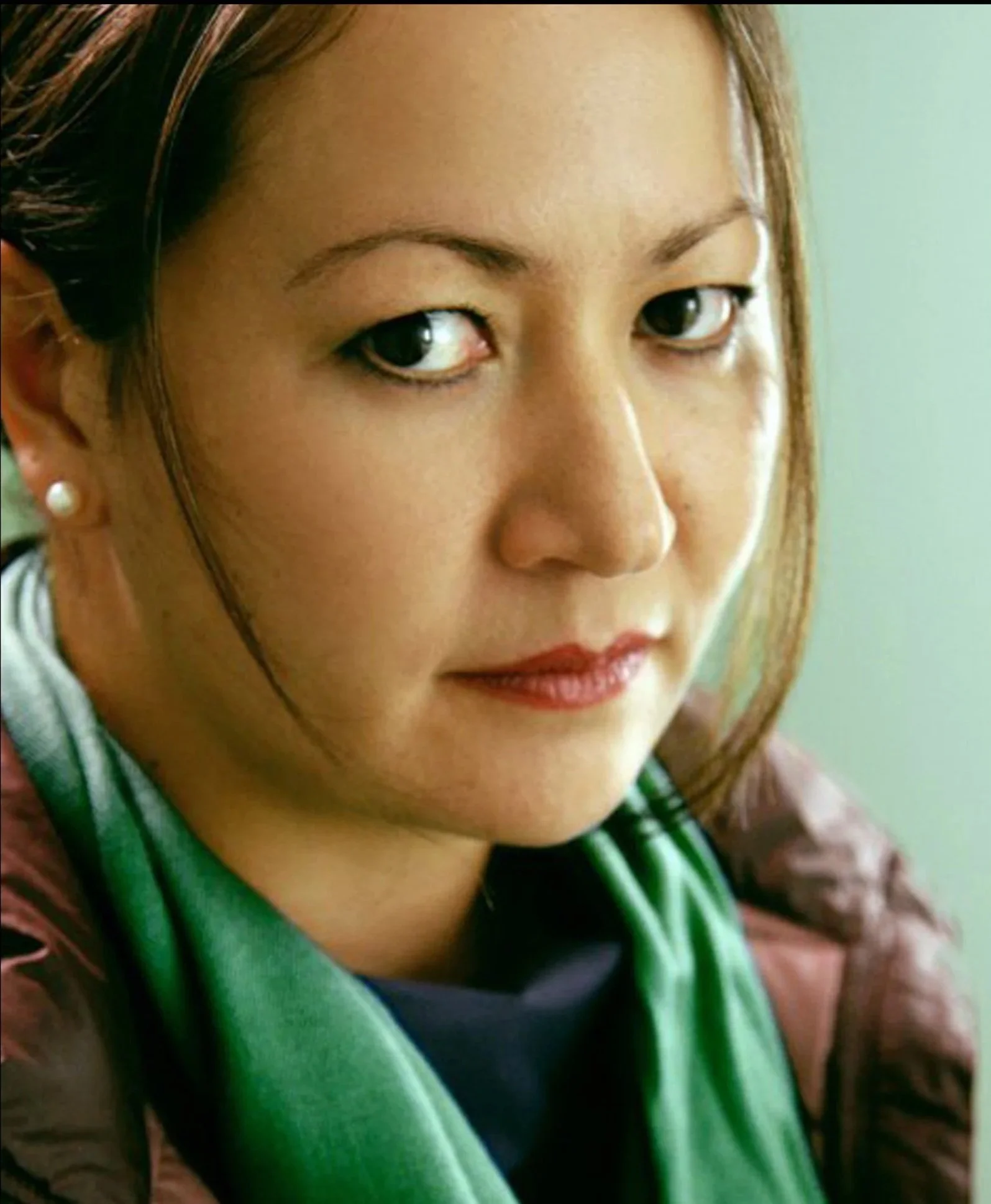 Close-up of a woman with brown hair, wearing pearl earrings, with a stethoscope around her neck, looking at the camera.
