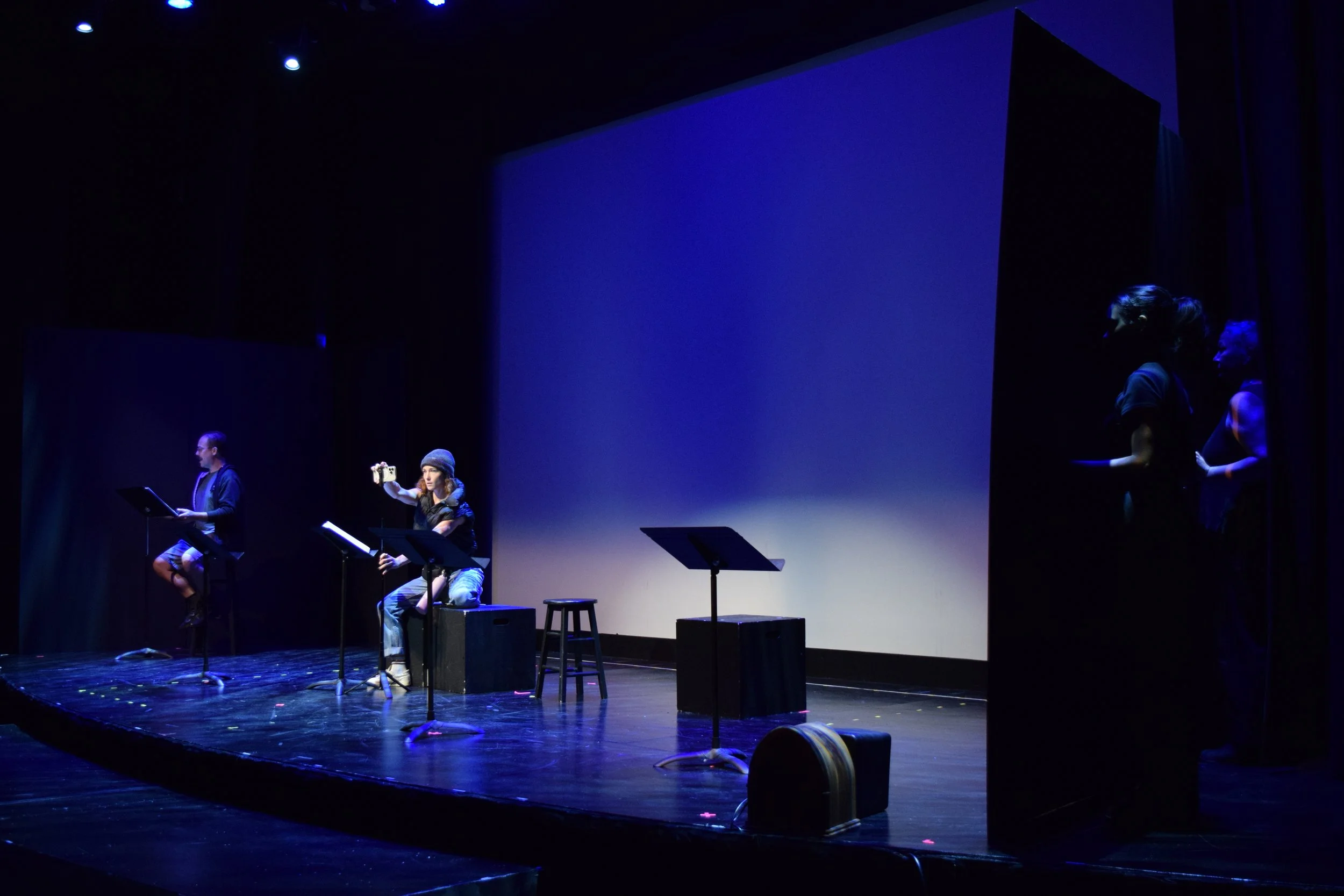 Four performers on a dark stage, seated on stools and holding music sheets, with a large blank screen behind them. The stage is dimly lit with blue lighting.