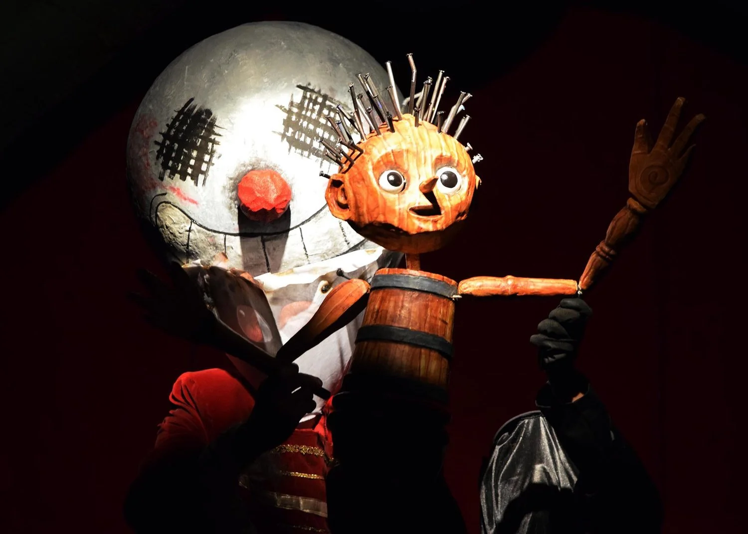 A puppeteered figure with a carved wooden face, large eyes, and a drum body, holding a staff, in front of a round moon-like prop with painted facial features.