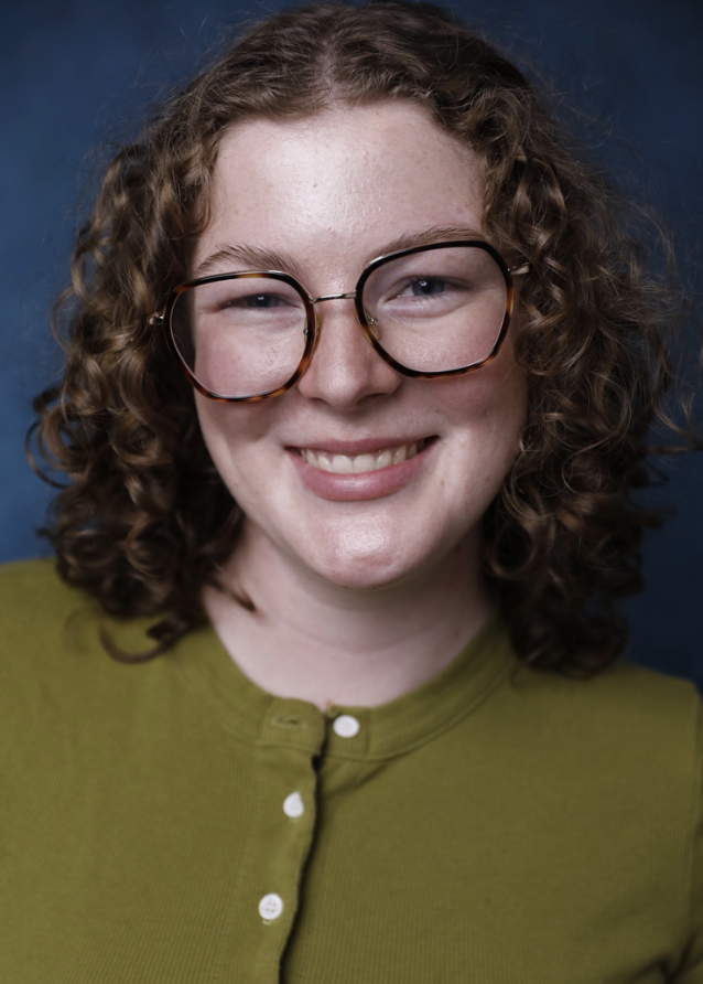 A young woman with curly red hair wearing glasses and a green shirt, smiling at the camera, with a dark blue background.