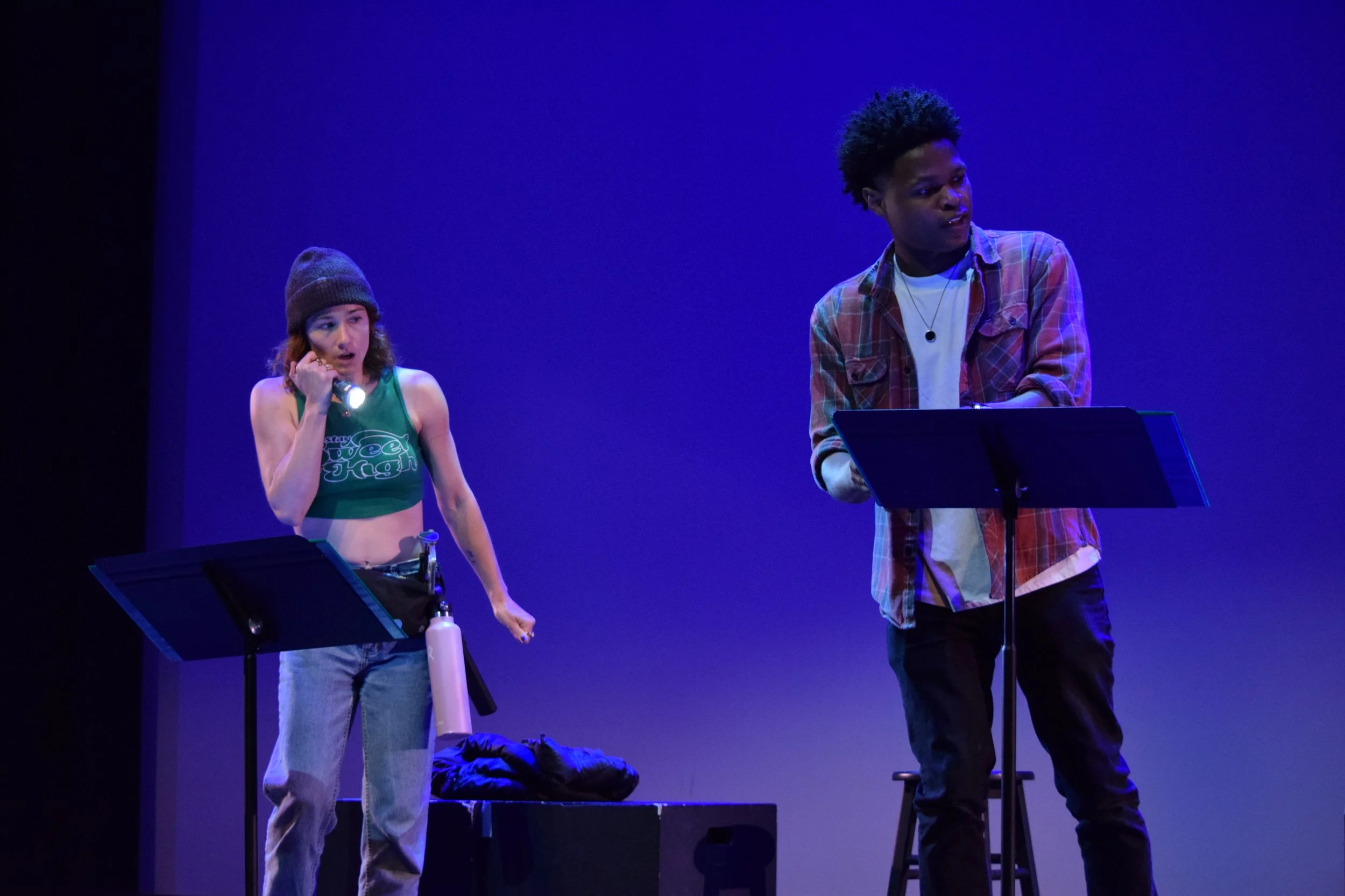 Two people on stage, one with reddish-brown hair wearing a gray beanie, green tank top, and gray pants holding a flashlight; the other with curly black hair wearing a plaid shirt over a white t-shirt, standing behind a music stand. The background is 