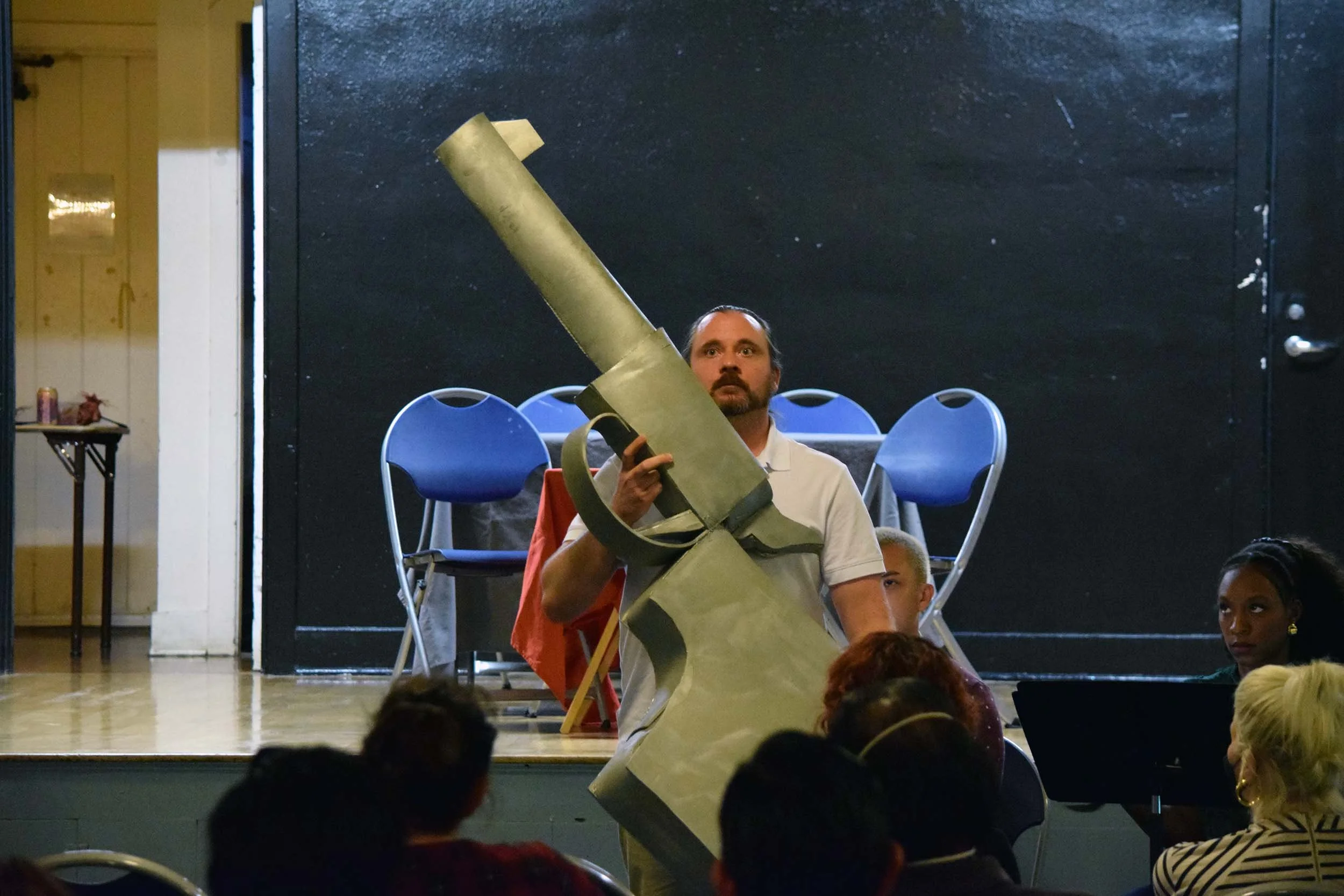 A man with a beard and white shirt holding a large piece of foam or cardboard, with children and adults seated on the floor in a community hall or indoor space.