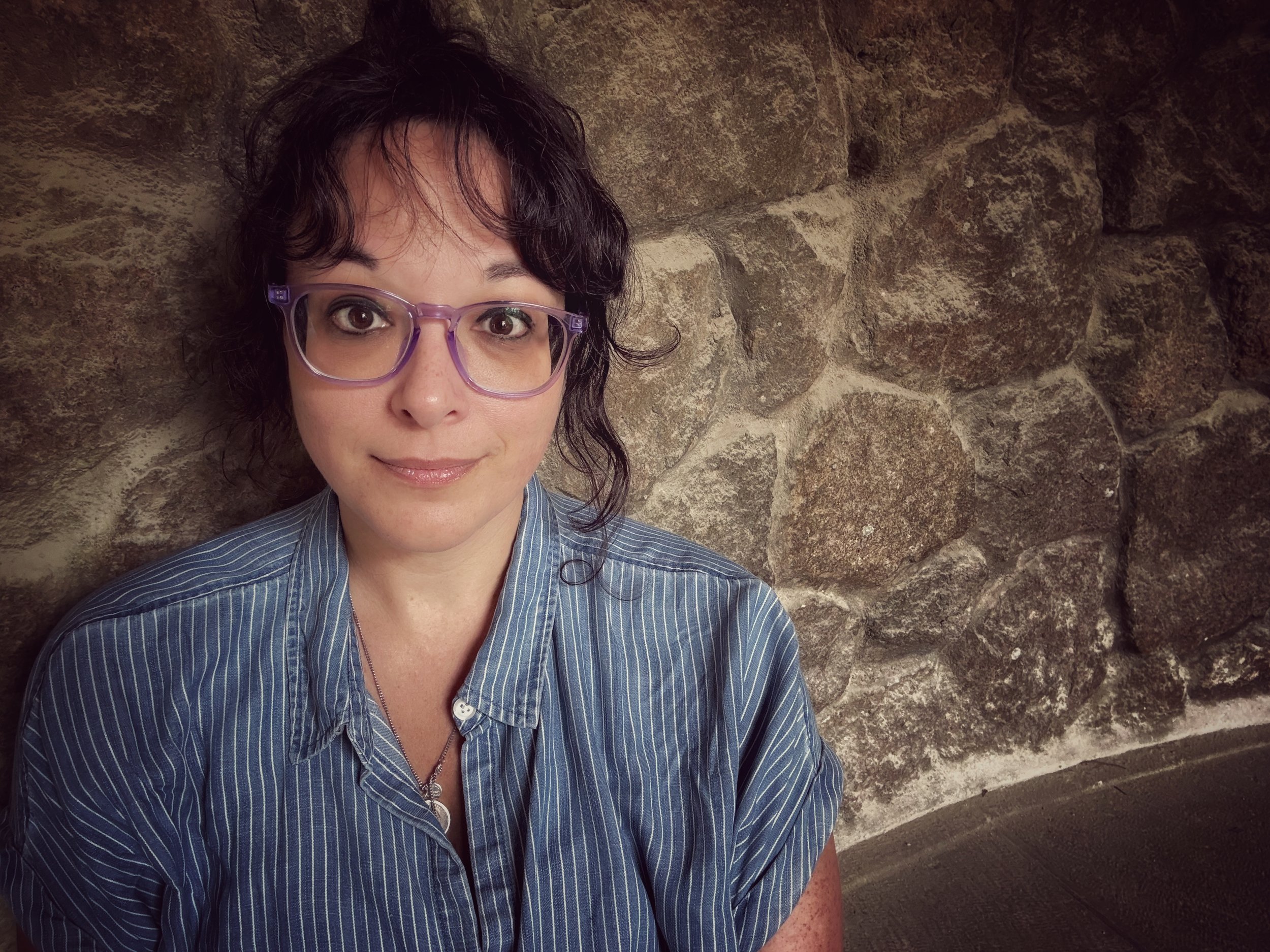 A woman with curly dark hair and glasses taking a selfie against a stone wall.