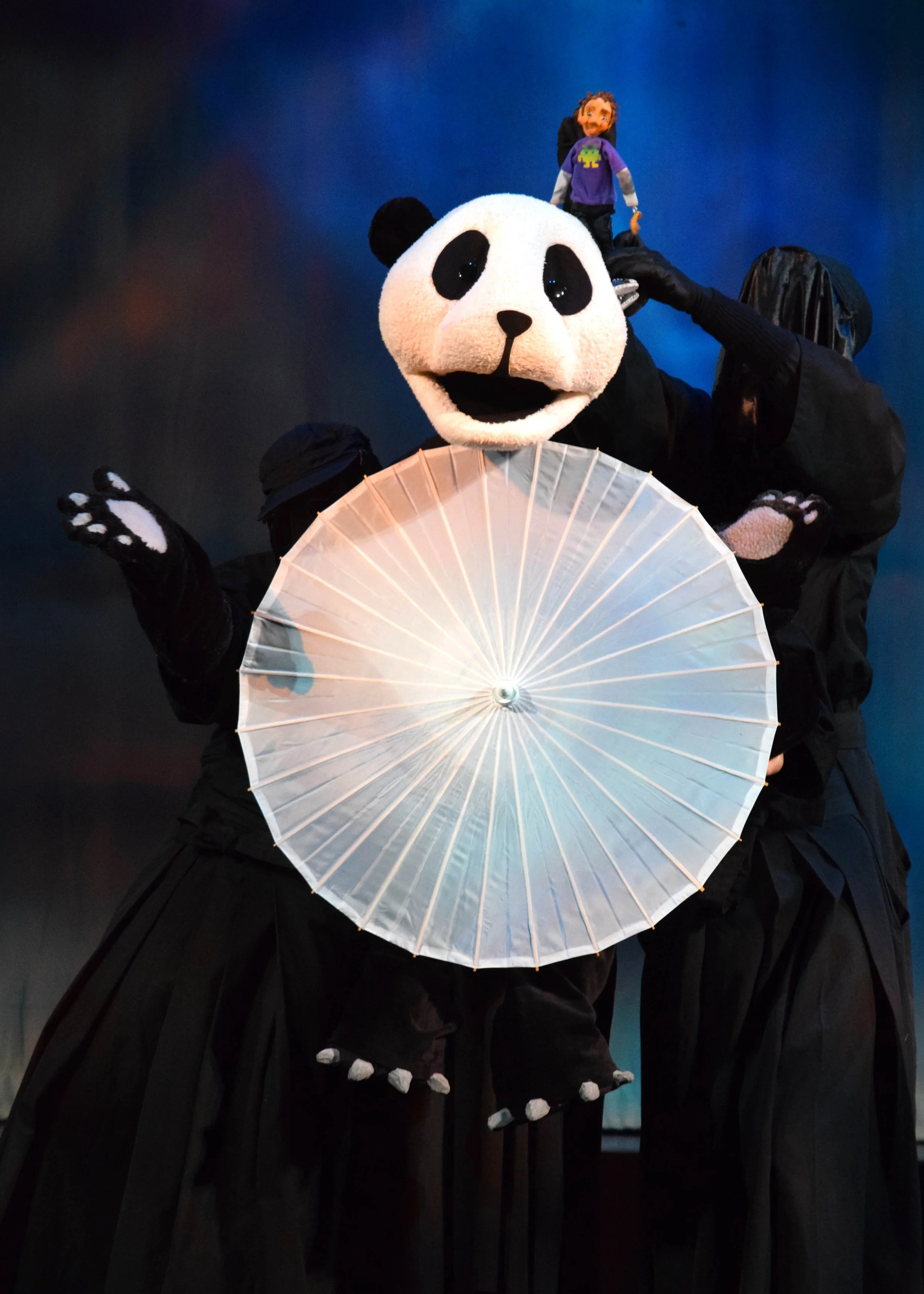 A person dressed in a panda costume holding a white parasol, with puppets of a child and an adult on their shoulders against a dark background.