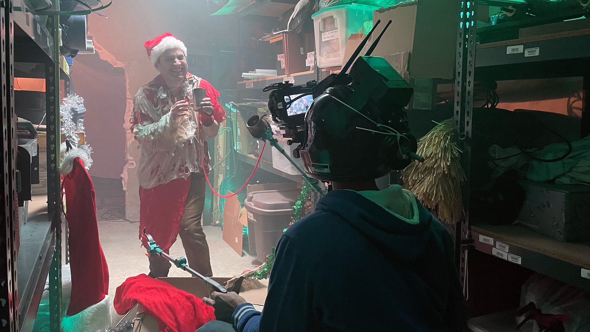 A person dressed in a Santa hat and holiday-themed clothing is celebrating with a microphone in a storage room decorated for Christmas. The room has shelves filled with boxes and equipment, with a video camera recording the scene. There are Christmas decorations like a red stocking, tinsel, and a small Christmas tree.