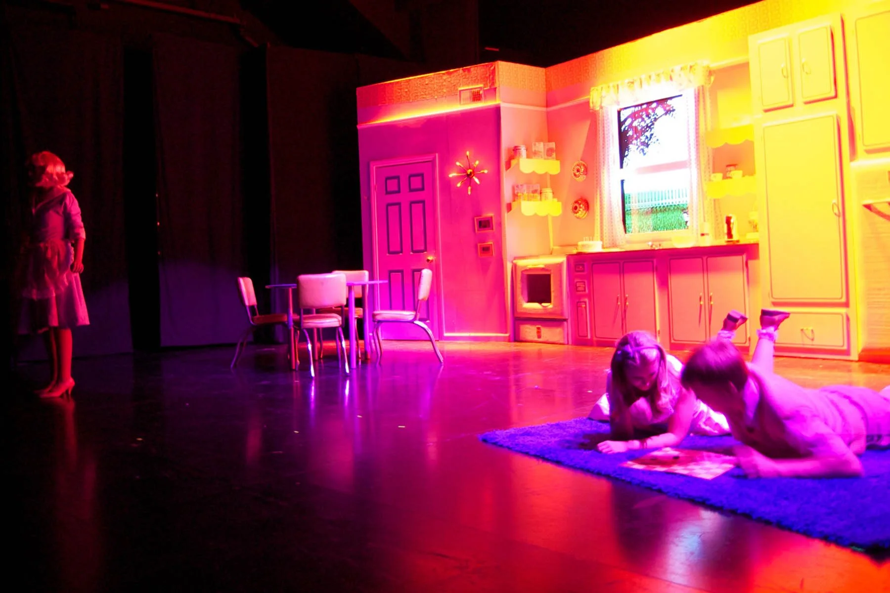 Children performing on a colorful stage decorated as a kitchen with a pink door, yellow cabinets, and a window. Two girls are lying on a rug, and a girl is standing on the left side.