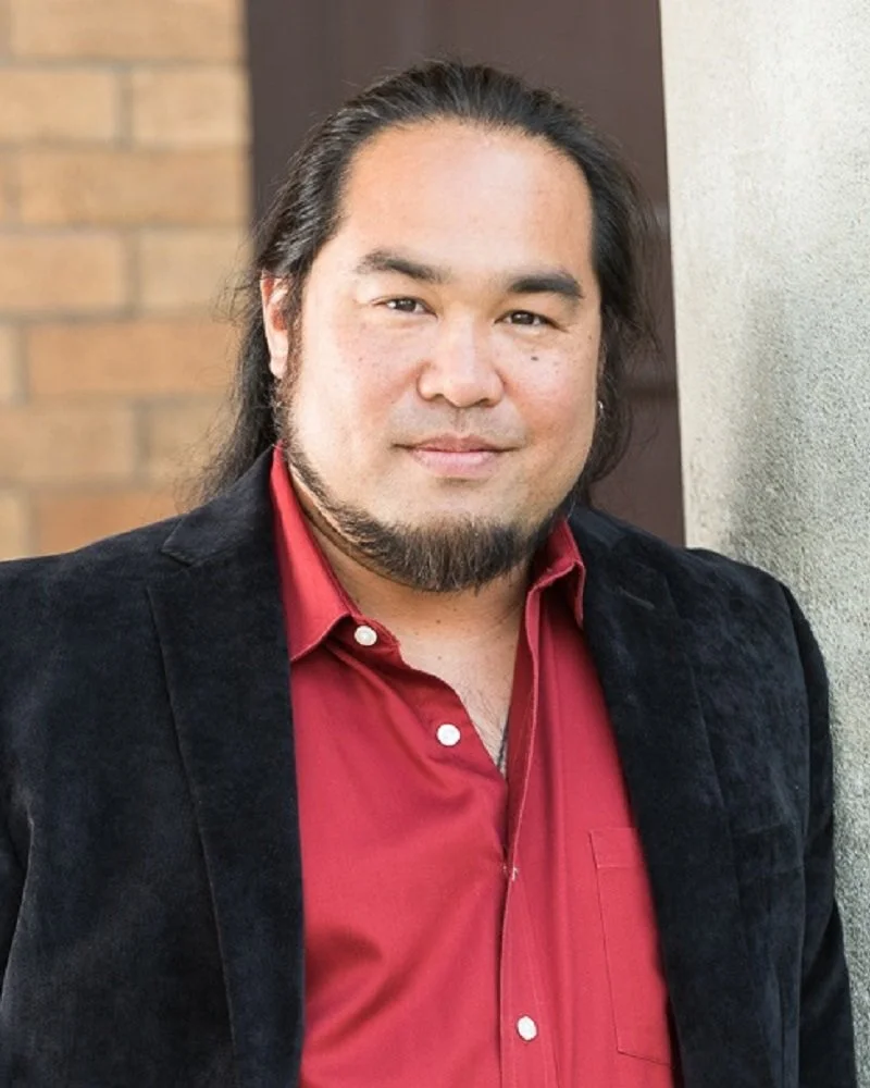 A man with long black hair, a beard, and a mustache, wearing a red shirt and black jacket, standing outdoors next to a concrete pillar with a brick wall in the background.