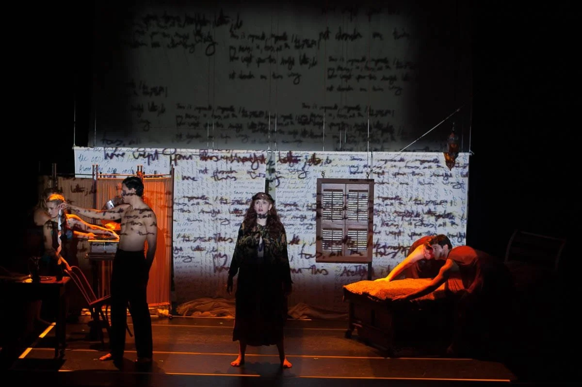 The scene depicts a theatrical stage with four actors under dim lighting and projected text background. One actor stands at center, while the others are engaged in various actions around the stage, including one leaning over a bed and another adjusting a screen or curtain.