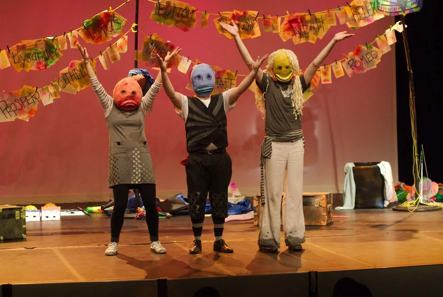 Three performers on stage wearing large, colorful masks with exaggerated facial features, raising their arms. Behind them, a backdrop with colorful flags and handwritten words and names, suggesting a theatrical performance.