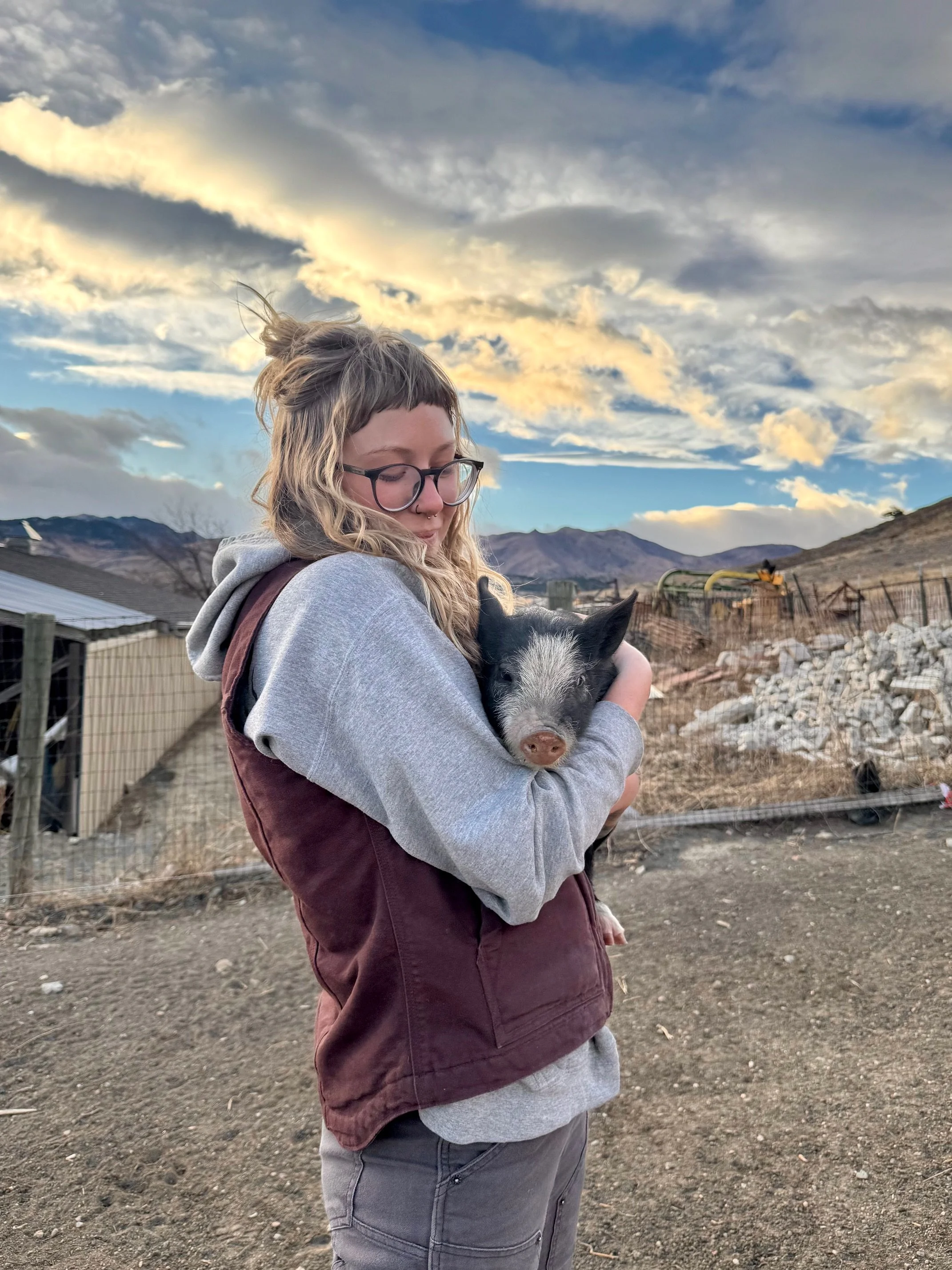 A woman with glasses and wavy hair holding a piglet with black and white fur against a backdrop of mountains, cloudy sky, and rural farmland.