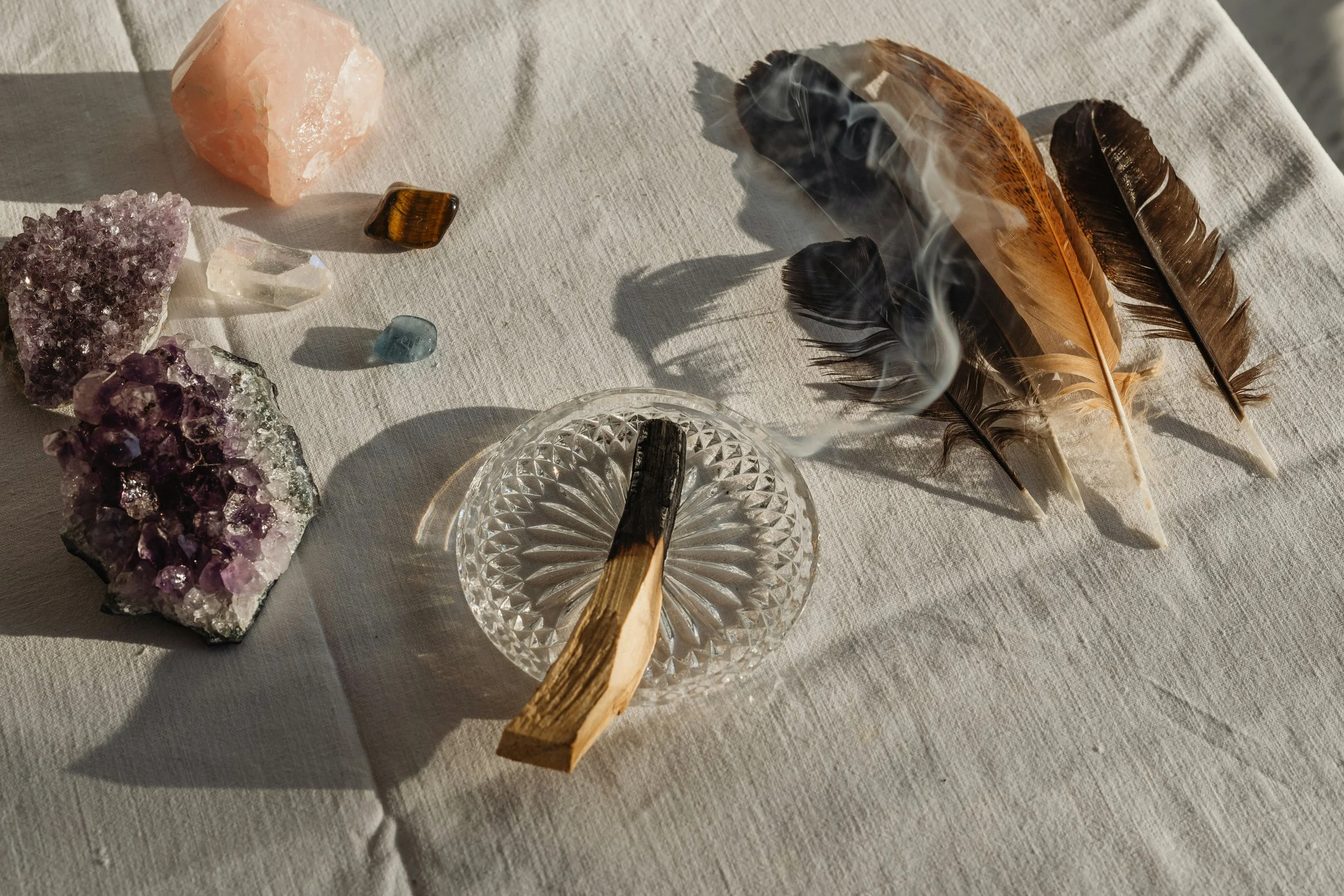 Arrangement of crystals, feathers, and a smoldering matchstick on a light cloth surface.