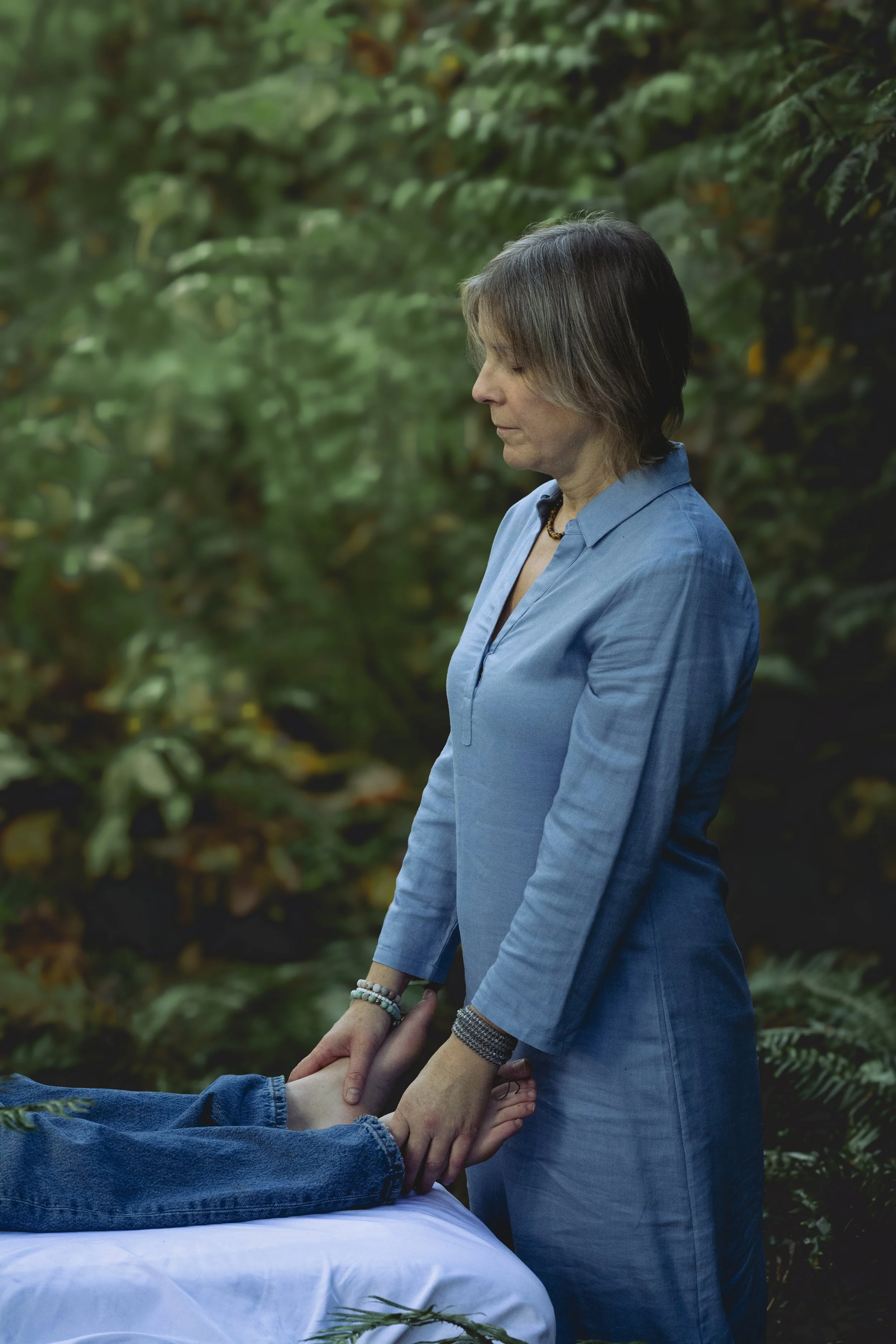 A woman in a blue dress giving a hand massage to a person lying on a white cloth in a forest setting.