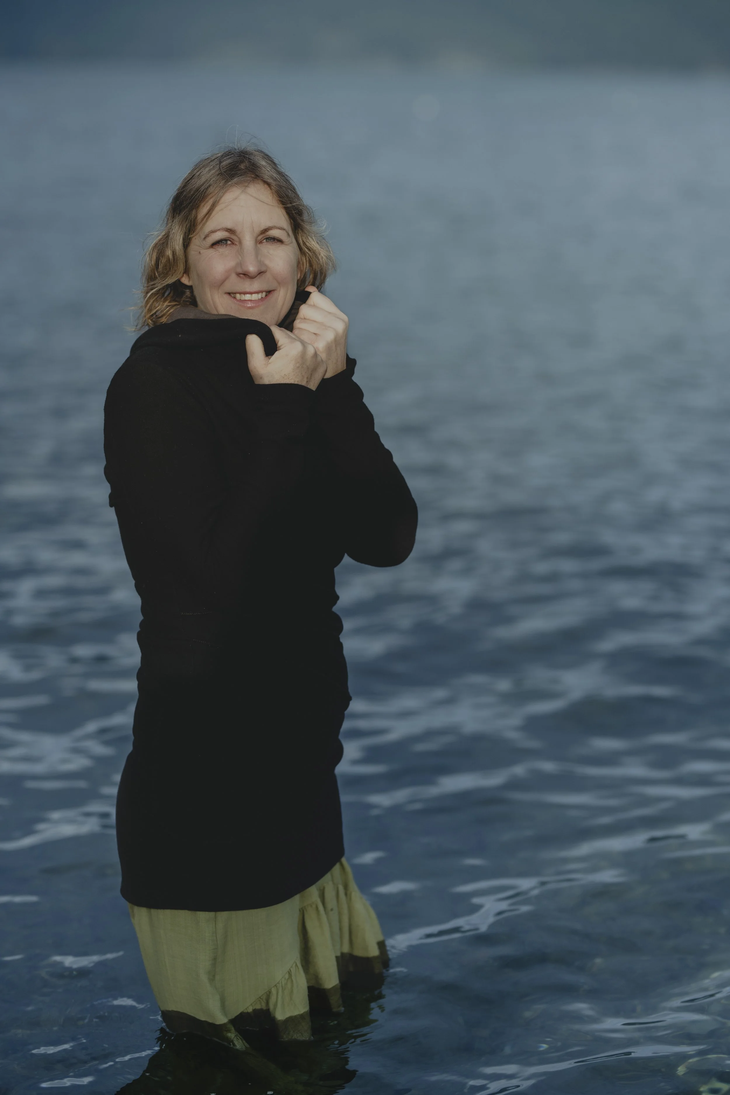 A woman standing in water near the shore, smiling, wearing a black jacket and yellow shorts.