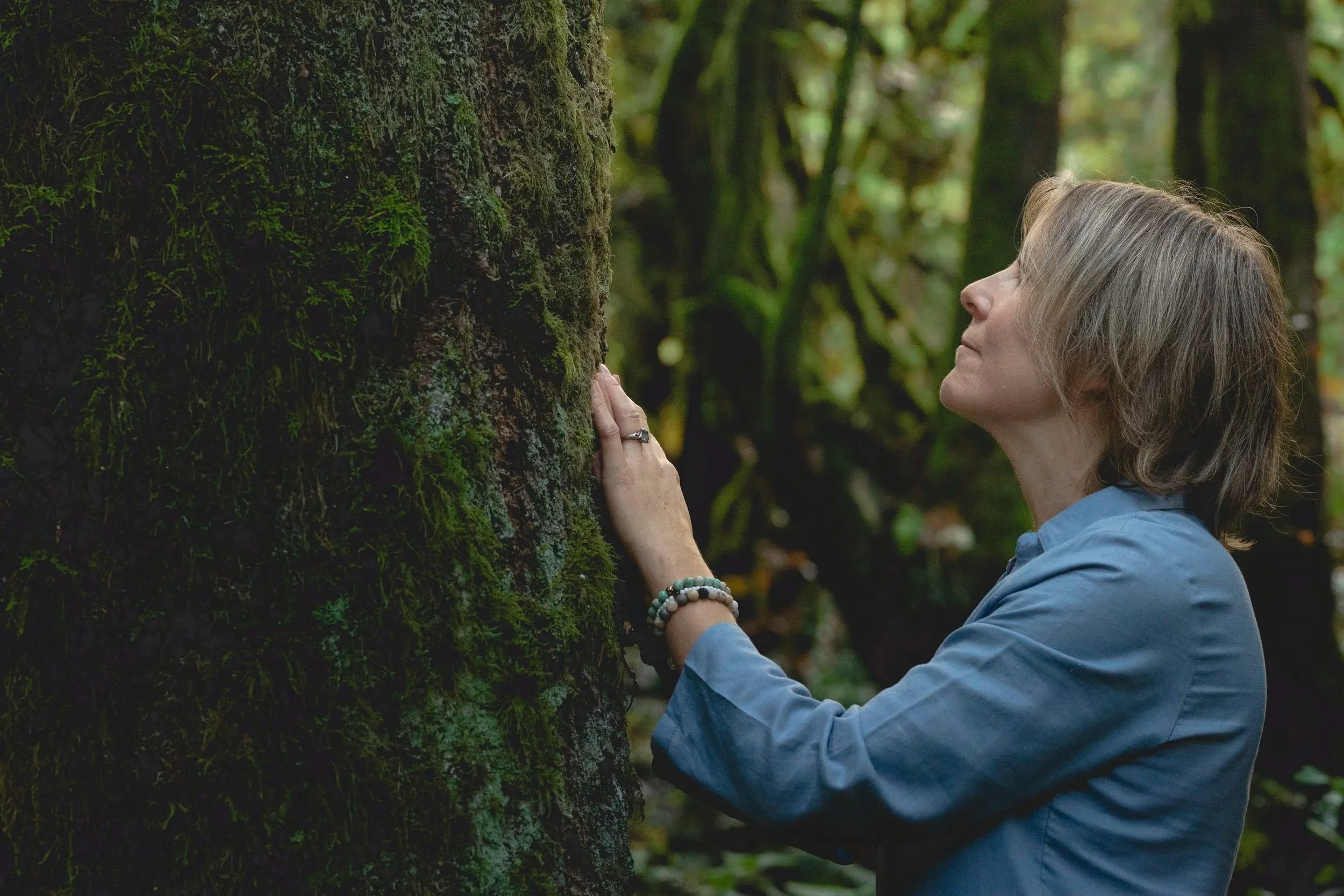 A woman with short brown hair wearing a blue shirt is standing next to a moss-covered tree in a forest, gently touching the tree trunk and looking at it with a serene expression.