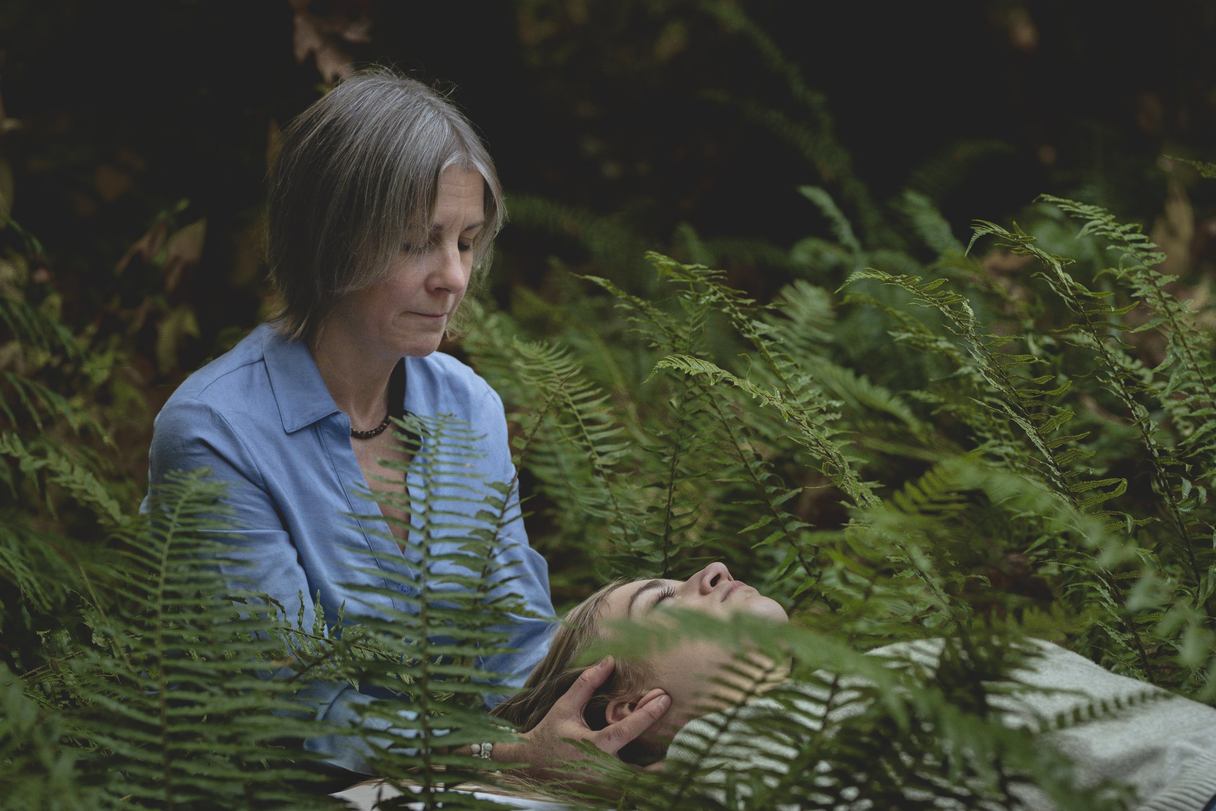 A woman in a blue shirt tending to a young girl lying among green ferns in a forest.