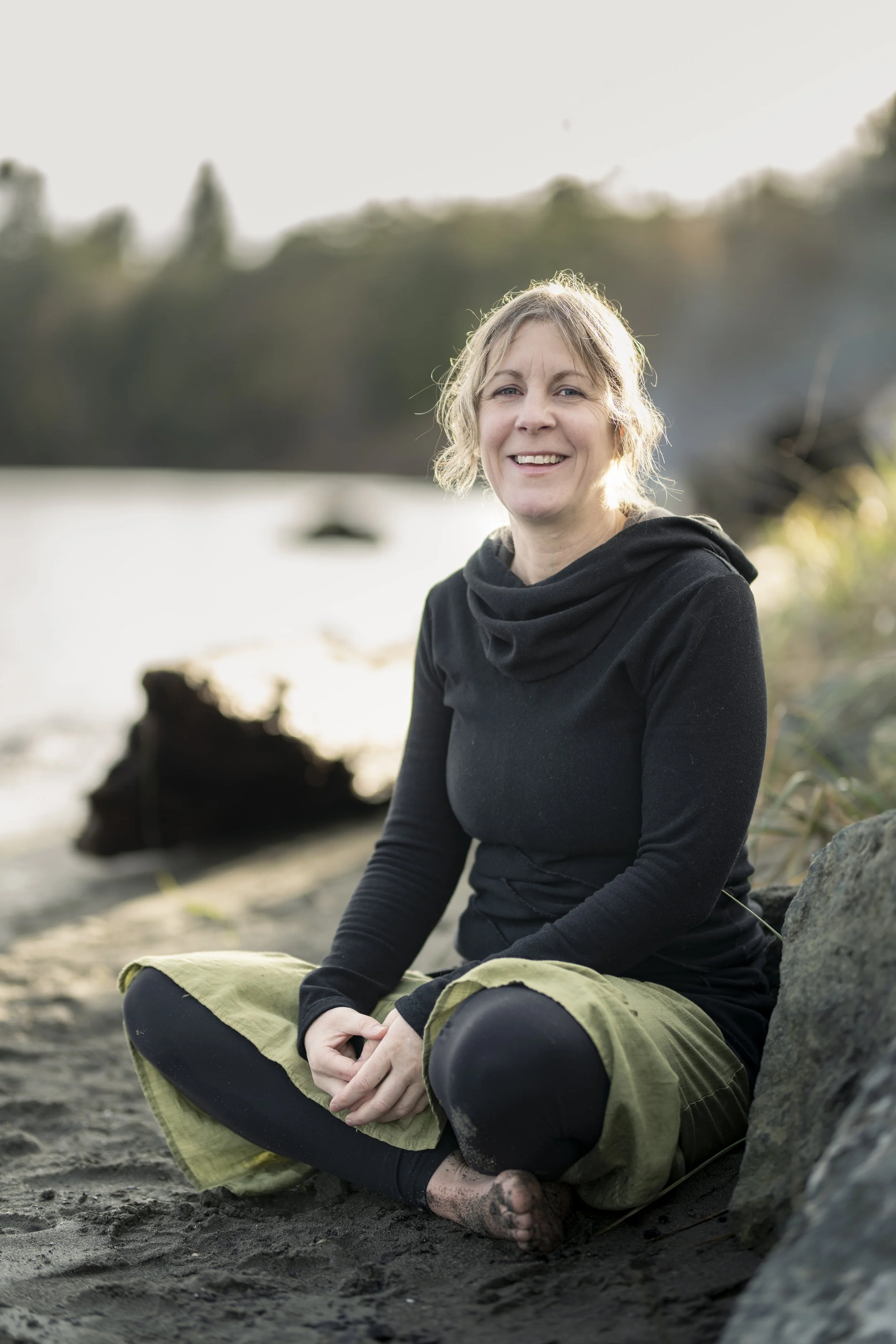 A woman sitting on a sandy beach near rocks, smiling at the camera with a blurred background of water and trees.
