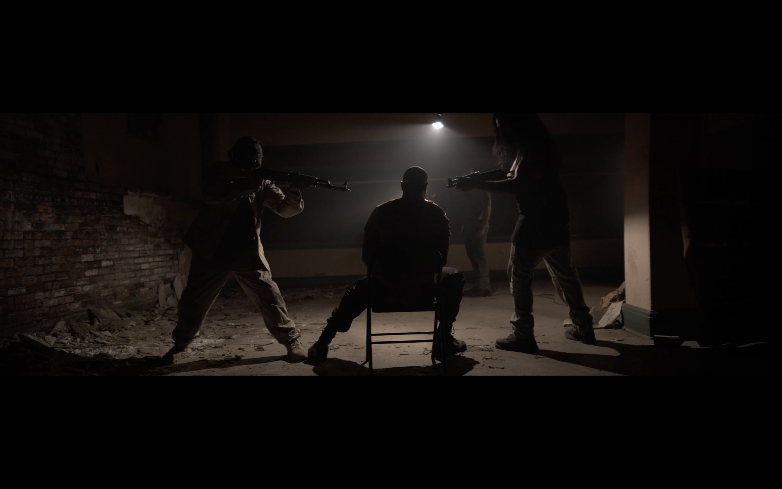 Silhouettes of three people holding guns and aiming at a seated person in a dark, dilapidated room with a brick wall and rubble, creating a tense scene.