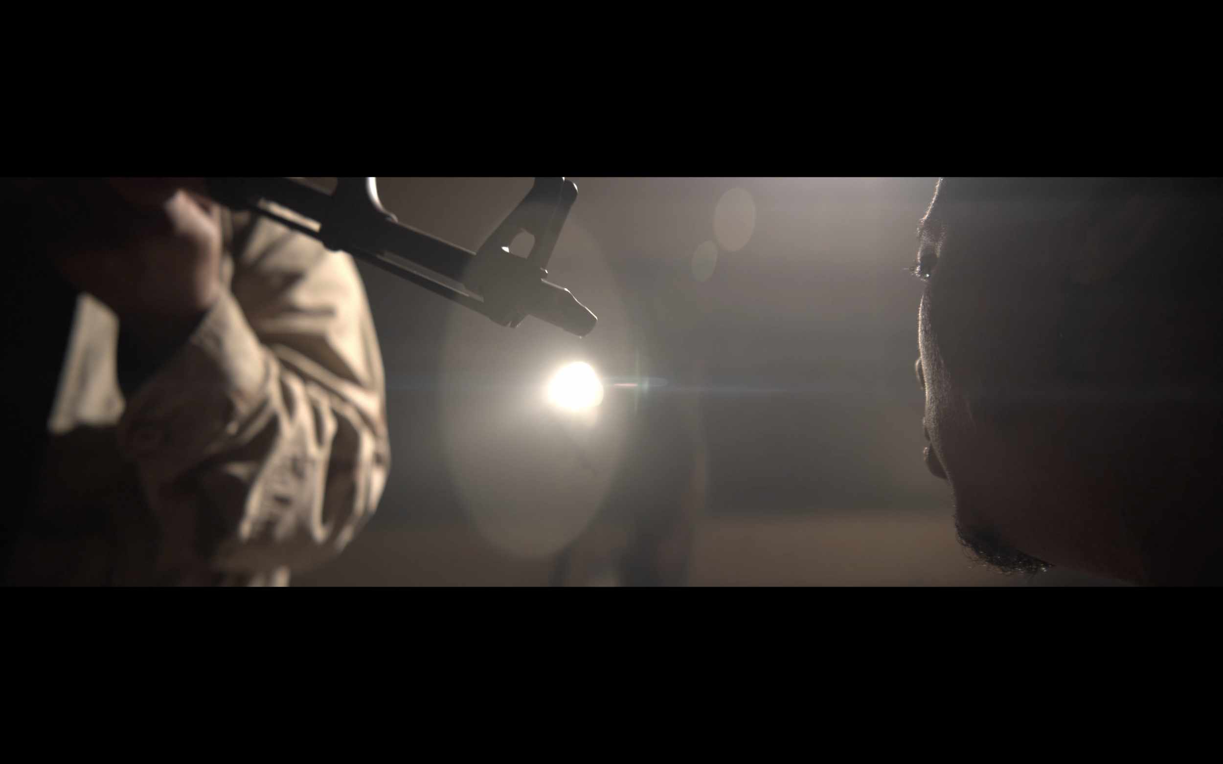 A person on the right side has a serious face, with a shadowed profile, and a beard. The person on the left side, only partially visible, is holding a pistol pointed at the person on the right. The background is dark, with bright light source creating lens flare effects, emphasizing the tension in the scene.