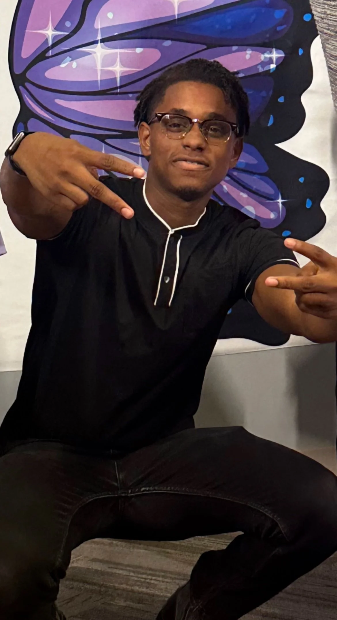A young man with glasses and short dreadlocks making a peace sign with his left hand and pointing at himself with his right hand, sitting in a room with a colorful butterfly mural painted on the wall behind him.