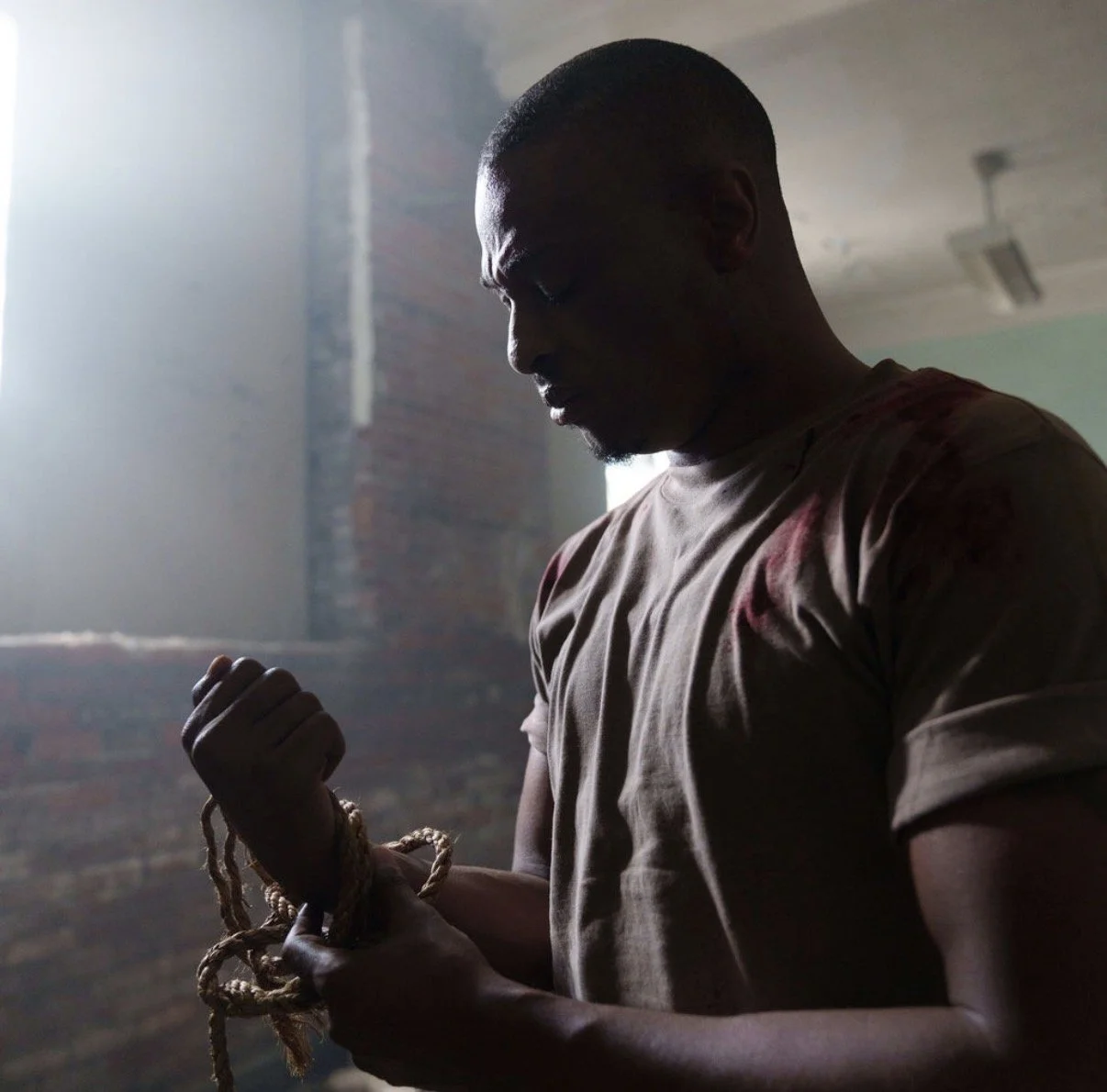 A young man in a dimly lit room holding a rope with a knot, looking down with a serious expression, in front of a brick wall and a large window.