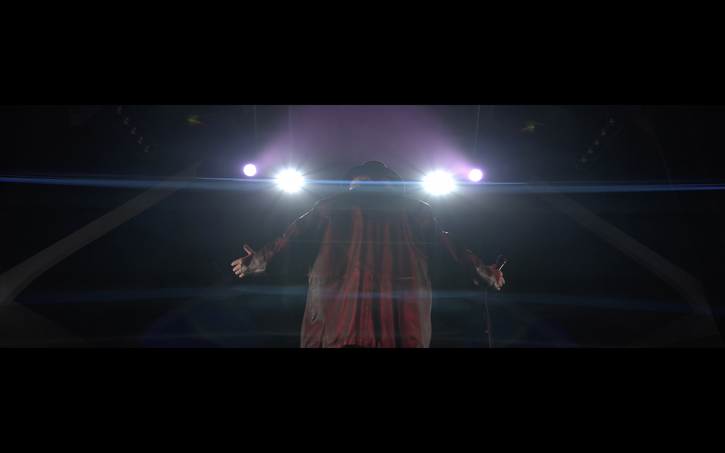 A person standing on stage with arms outstretched, facing four bright stage lights, wearing a hat and a striped shirt, backlit creating a silhouette effect.