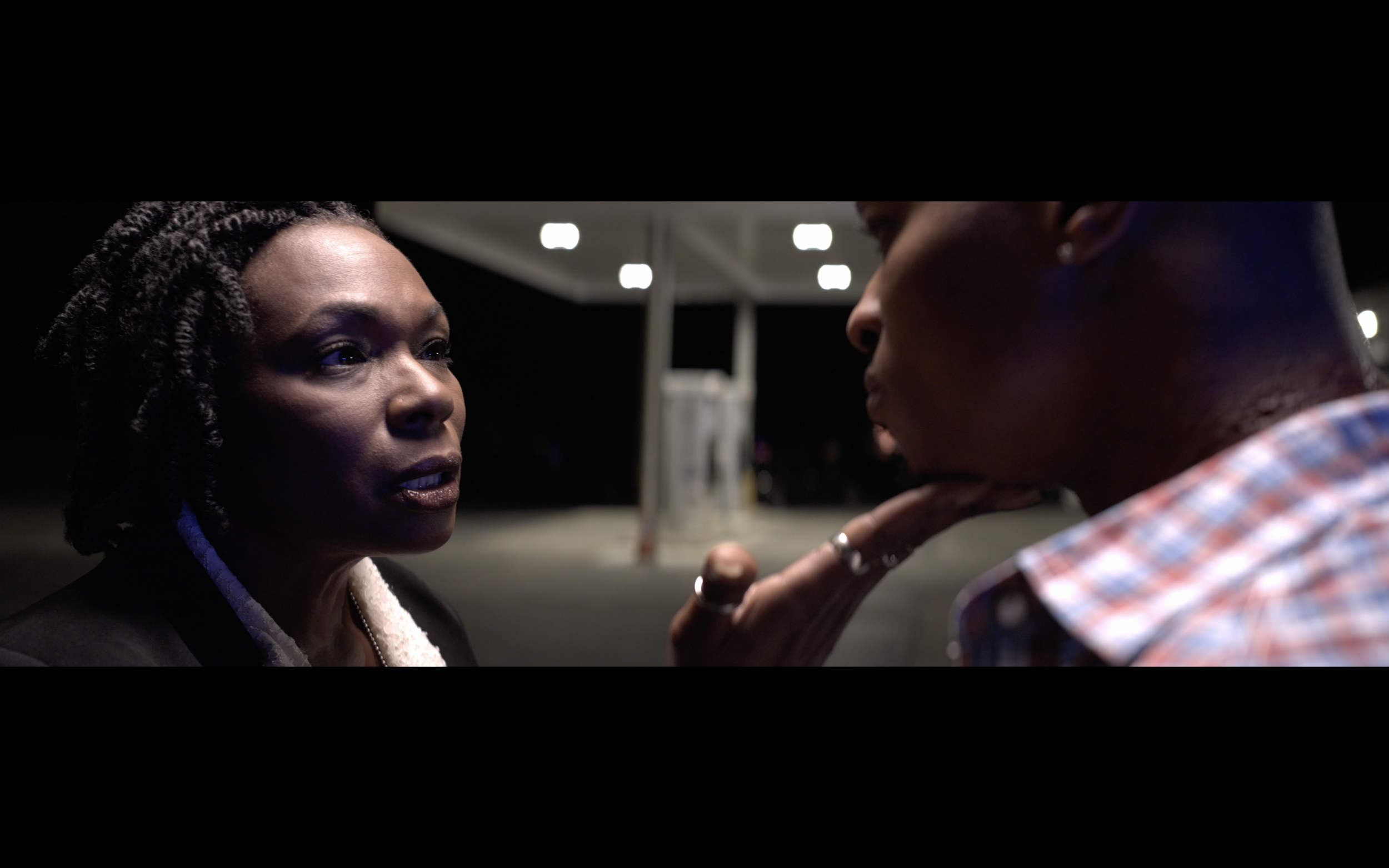 A woman with dark braided hair and a man with dark skin, short hair, and a checkered shirt are engaged in a close, intense conversation outdoors at night with bright lights in the background.