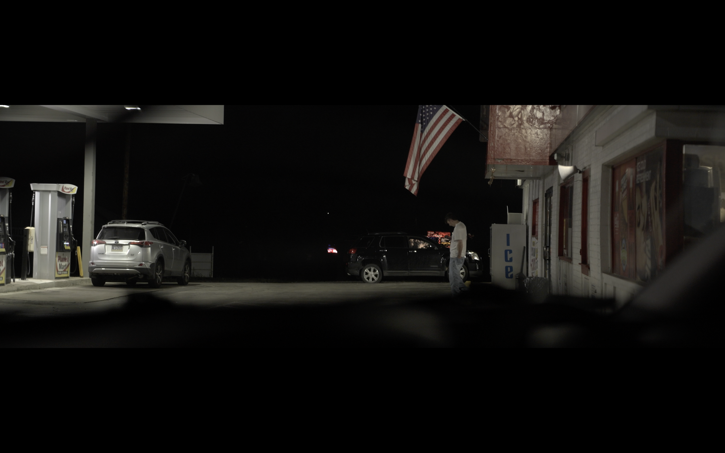 Night scene at a gas station with an American flag hanging outside. Several parked cars and a person standing near a black vehicle are visible. The gas station building has a white exterior with red accents and a vending machine nearby.