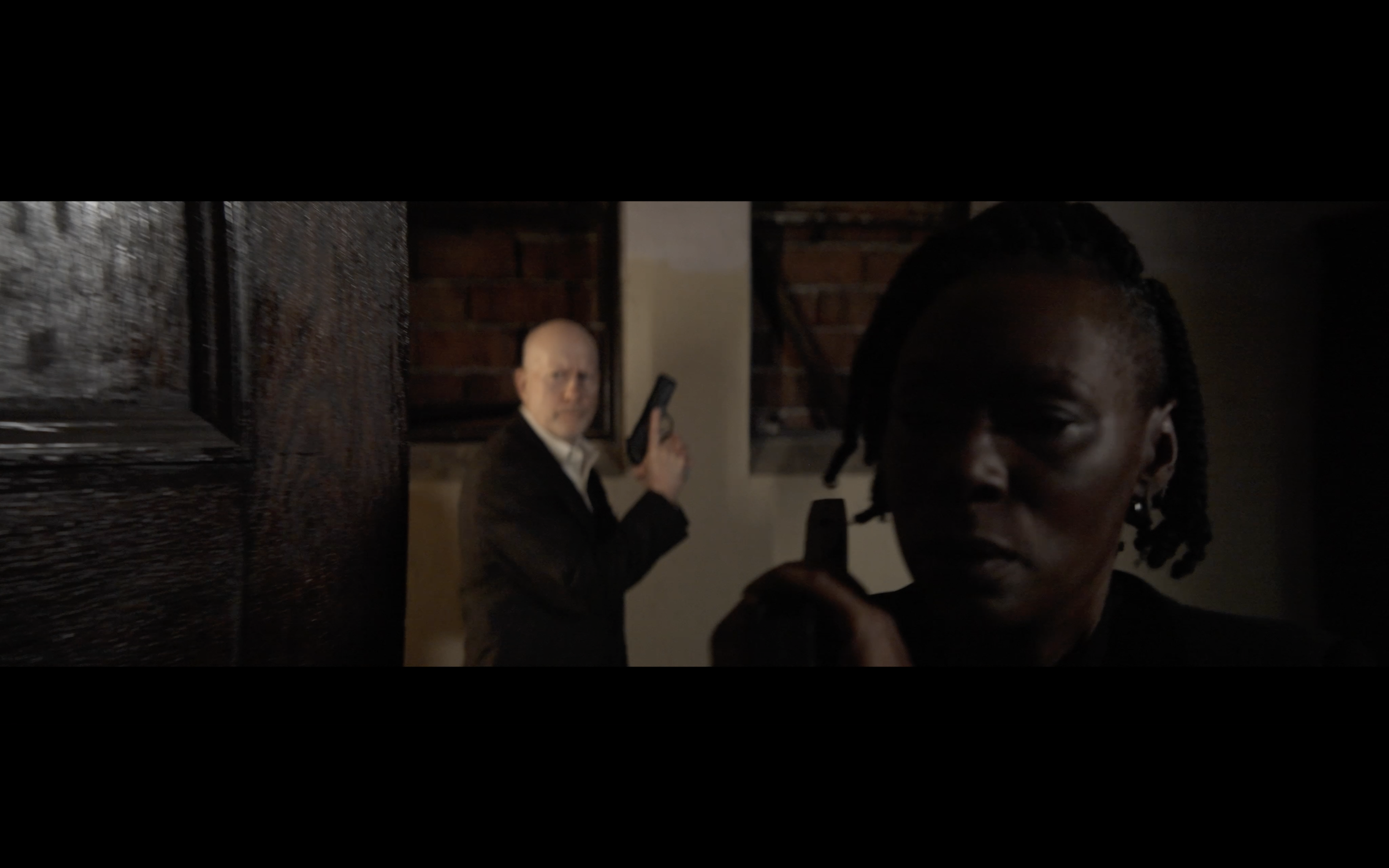 A woman with dreadlocks taking a selfie and a man with a bald head and white shirt holding a gun in a room with brick walls.