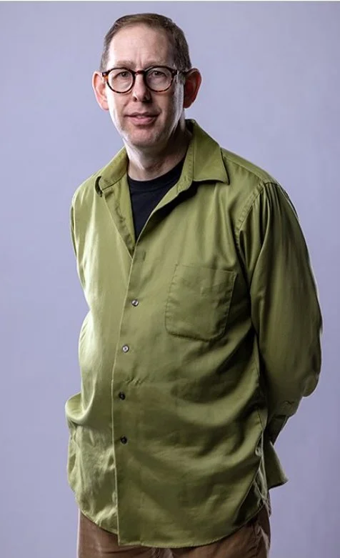 A man with glasses wearing a green button-up shirt over a black t-shirt, standing against a gray background.
