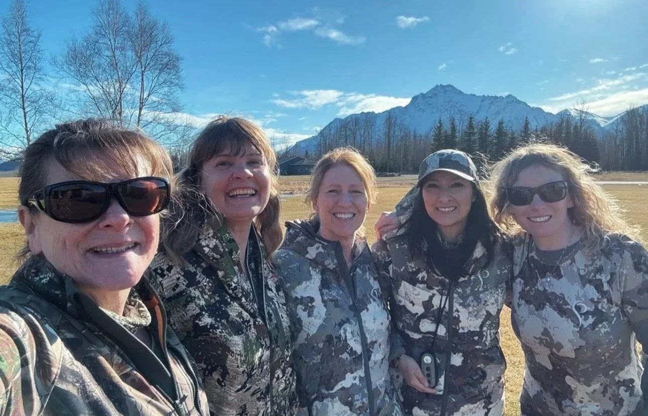 Five women in camouflage clothing smiling outdoors with mountains, trees, and a partly cloudy sky in the background.
