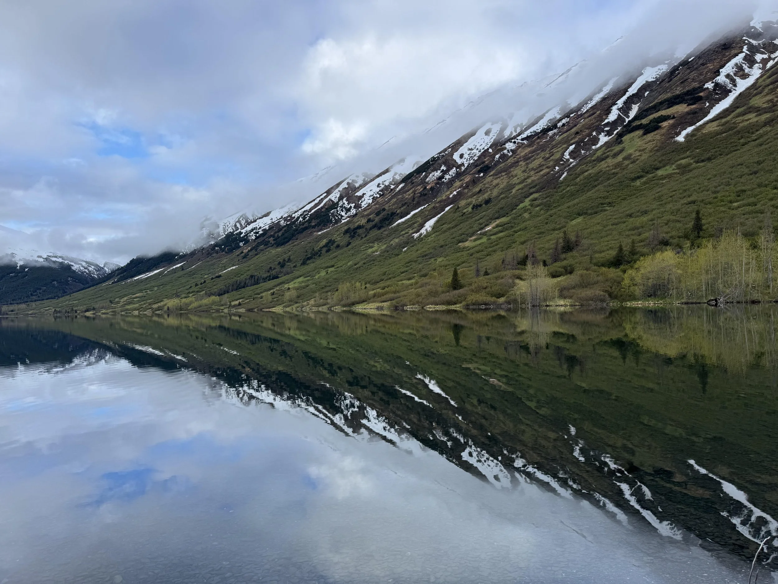 Snow-capped mountains with green slopes and trees reflected in a calm lake under a partly cloudy sky.