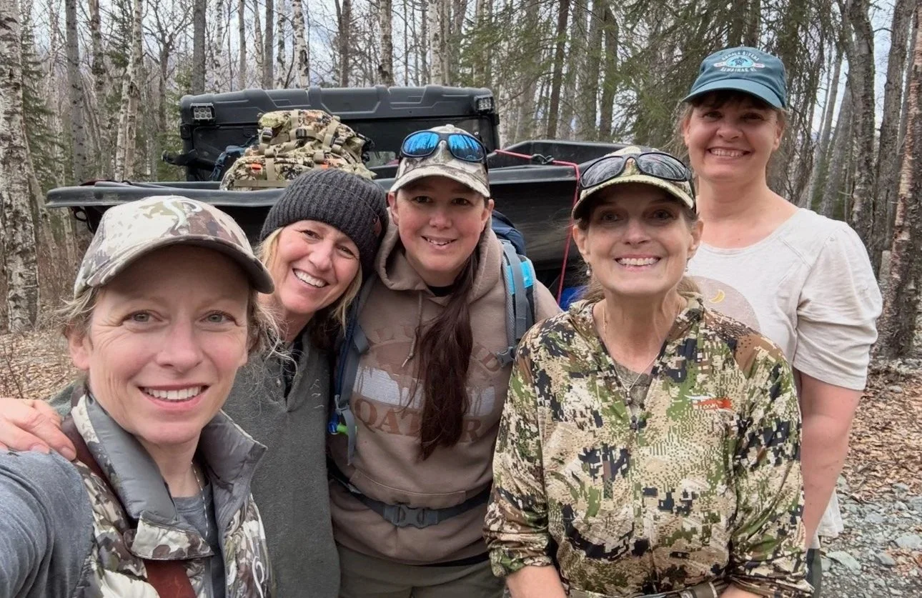 Group of five women outdoors in a wooded area, smiling for a selfie, with backpacks and outdoor clothing, suggesting they are on a hiking adventure.