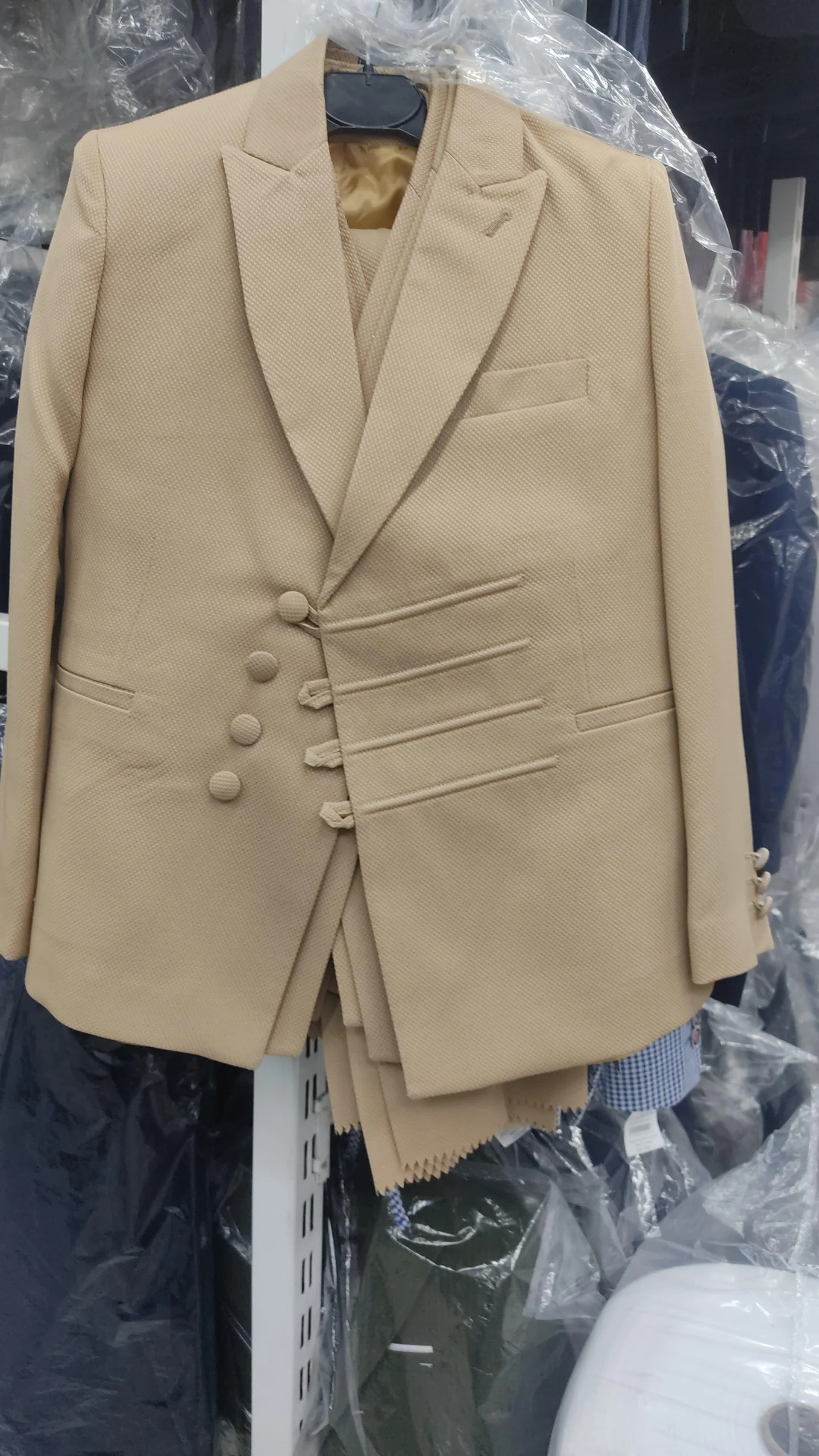 A beige men's suit jacket with padded shoulders and matching trousers hanging on a black hanger. The suit jacket has four buttons and a notched lapel. The background contains plastic wrapping and other clothing items.