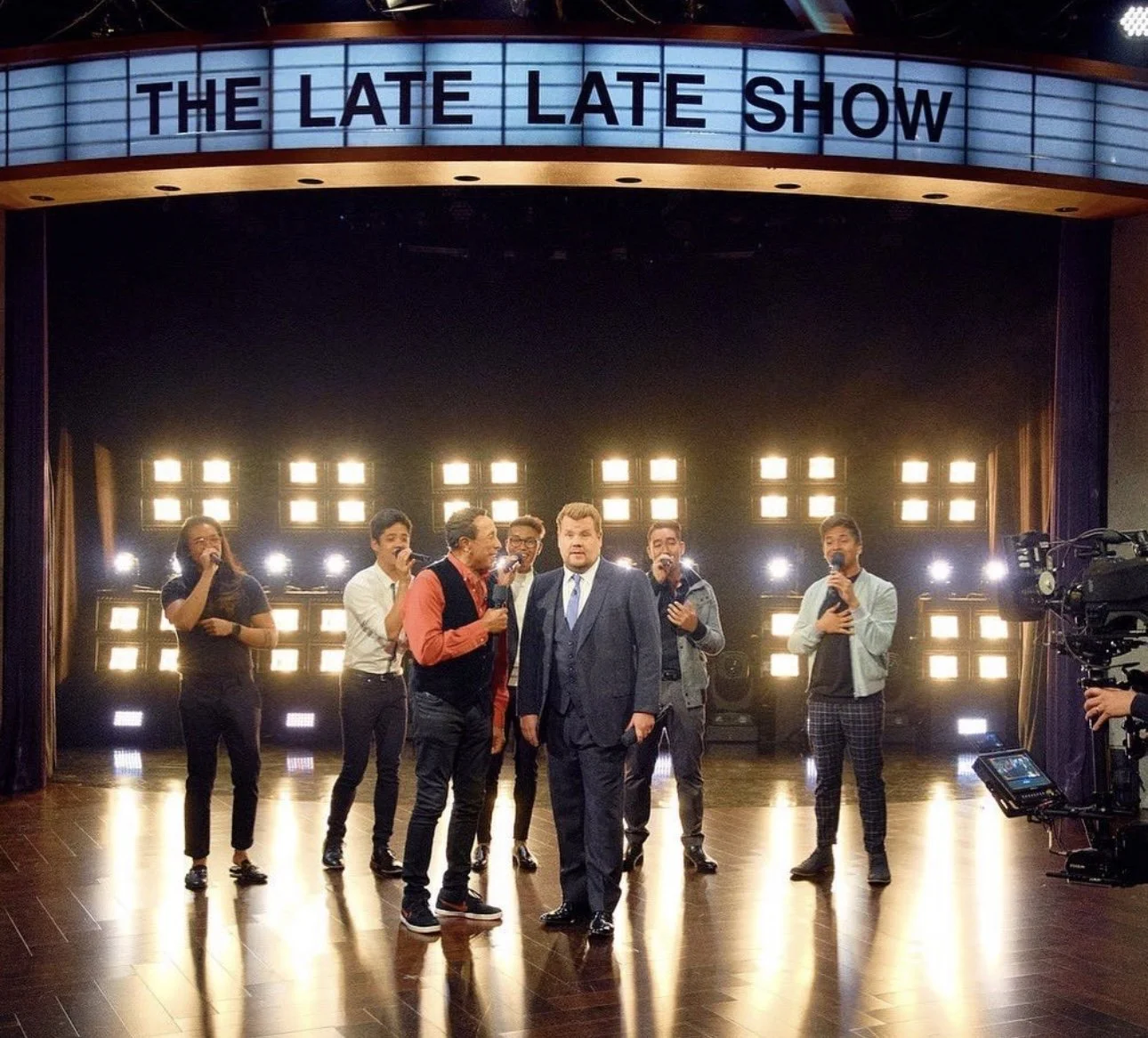 A group of seven people, including a man in a suit, singing on a television stage with bright lights behind them. The stage is associated with 'The Late Late Show' as indicated by the illuminated sign at the top.