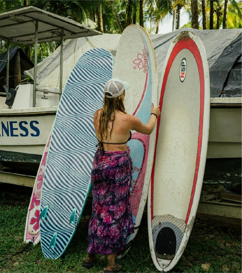 A woman with long hair wearing a baseball cap and a tropical skirt leans on and touches three surfboards propped against a boat. The surfboards are standing upright on grass, with a tropical background of palm trees.