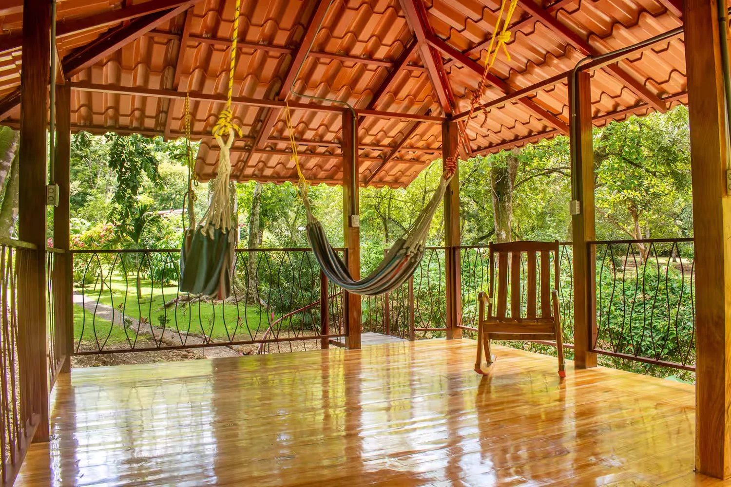 A wooden and tile porch with a hammock, a wooden chair, and a view of a lush green garden with trees and grass.
