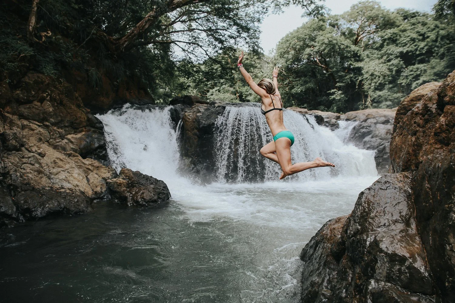 Woman jumping into a river in front of a waterfall surrounded by rocks and trees.