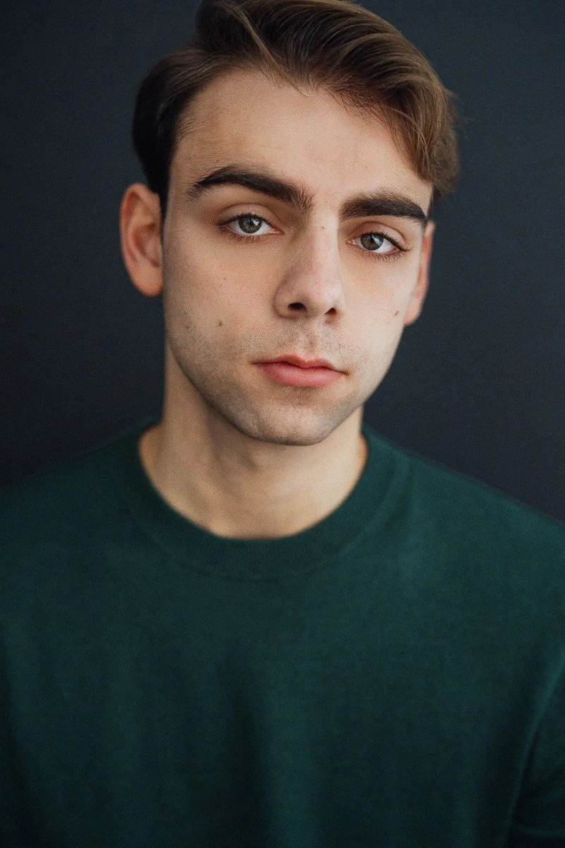 Close-up portrait of a young man with light skin, light brown hair, and blue eyes, wearing a dark green crewneck shirt, against a dark background.