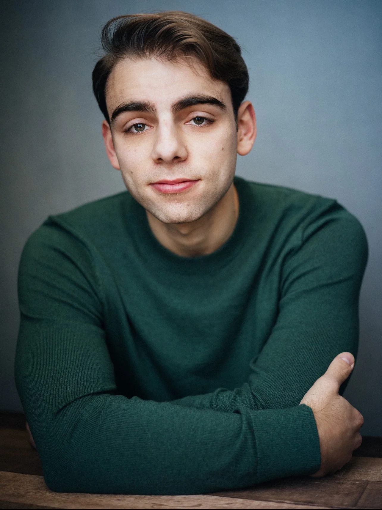 A young man with brown hair and light skin wearing a green long-sleeve shirt, sitting at a table with arms crossed, looking into the camera with a slight smile, against a plain gray background.