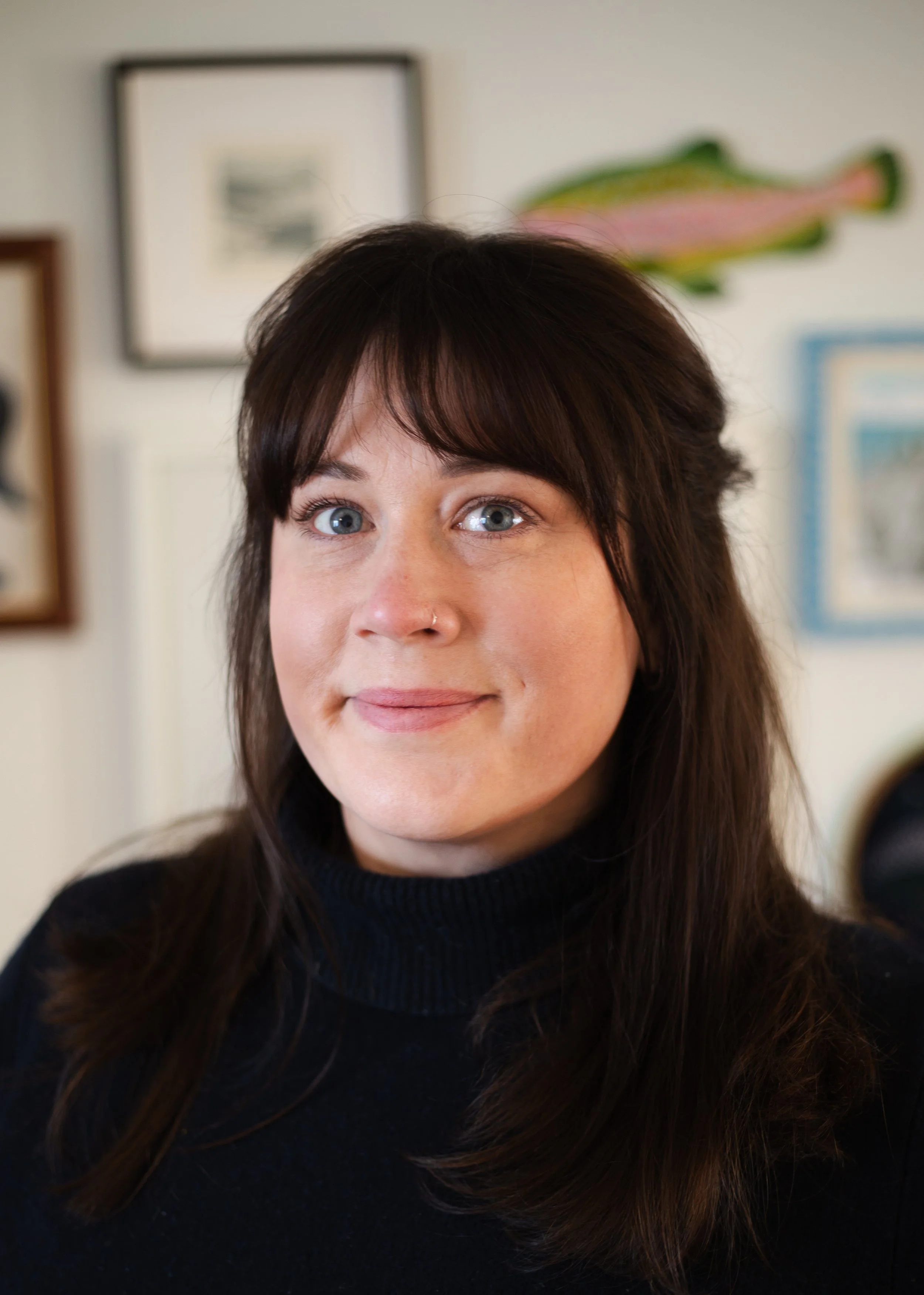 A woman with long dark hair and blue eyes, wearing a black turtleneck sweater, smiling indoors with framed artwork and a fish decoration in the background.