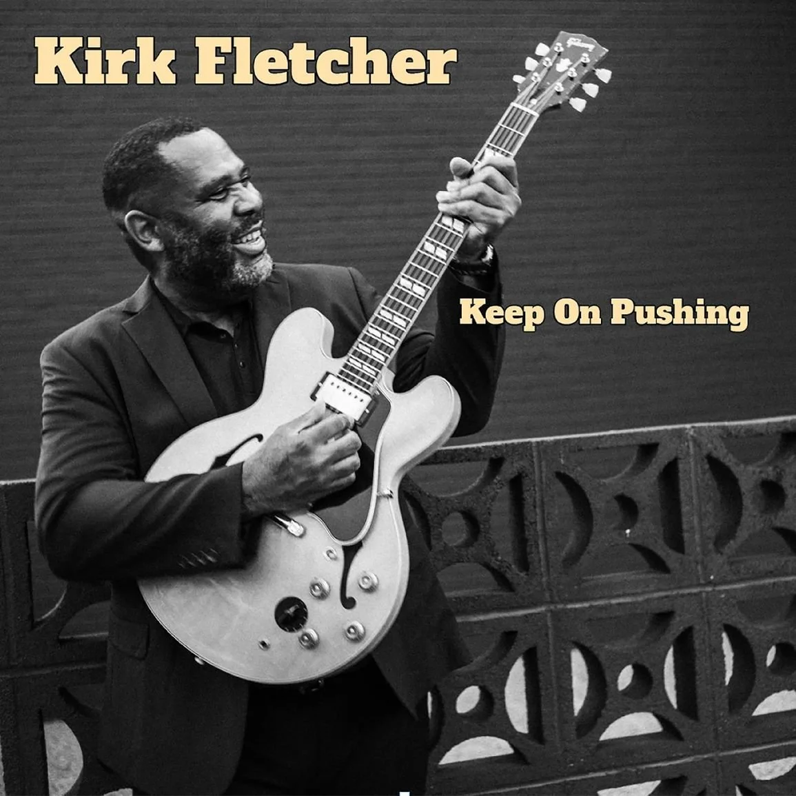 Schwarz-Weiß-Foto eines Mannes mit dunklem Anzug, der eine elektrische Gitarre spielt und vor einer gemusterten Wand steht. Oben links steht in gelber Schrift 'Kirk Fletcher' und rechts 'Keep On Pushing'.