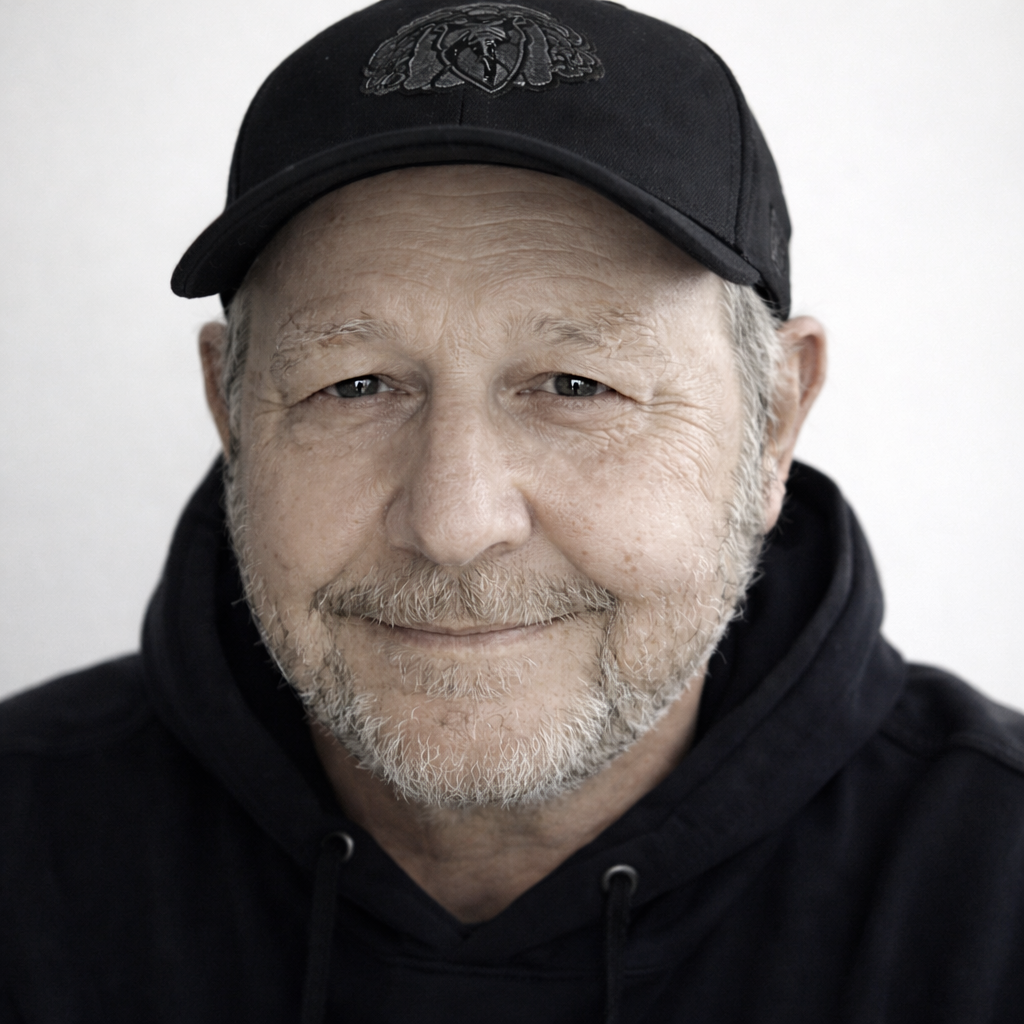 Nahaufnahme eines lächelnden älteren Mannes mit grauem Bart und Haaren, trägt eine schwarze Cap mit einem Emblem und einen schwarzen Hoodie.