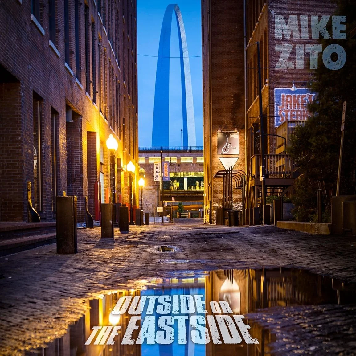 Blick durch eine urbane Gasse in St. Louis, mit dem Gateway Arch im Hintergrund, bei Dämmerung. Die Straße ist nass und reflektiert das Licht der Straßenlaternen und den Schriftzug "OUTSIDE OR THE EAST SIDE" auf der Wasserpfütze.