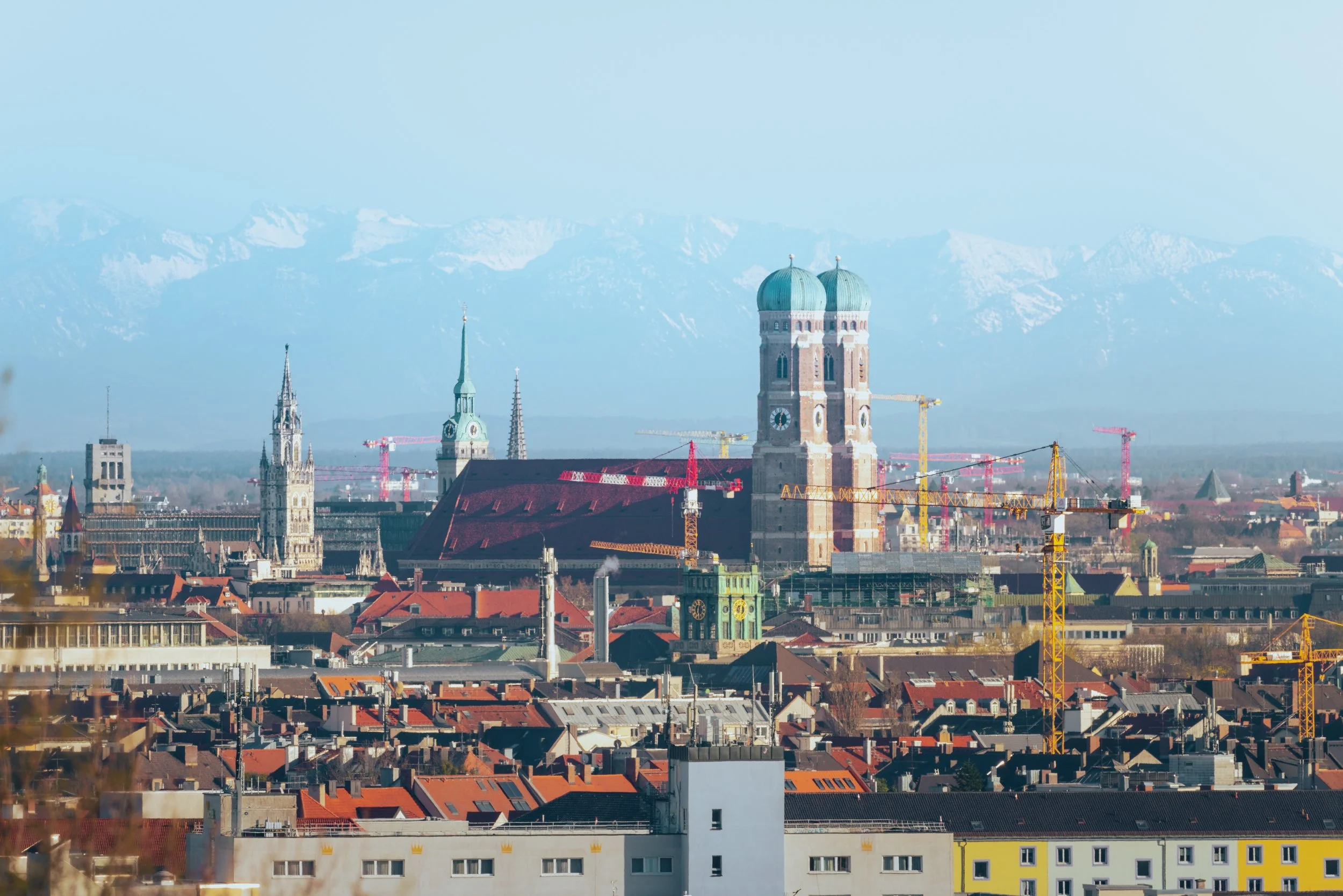 Stadt München mit Baukränen im Vordergrund, die Frauenkirche und andere historische Gebäude, Berge im Hintergrund.