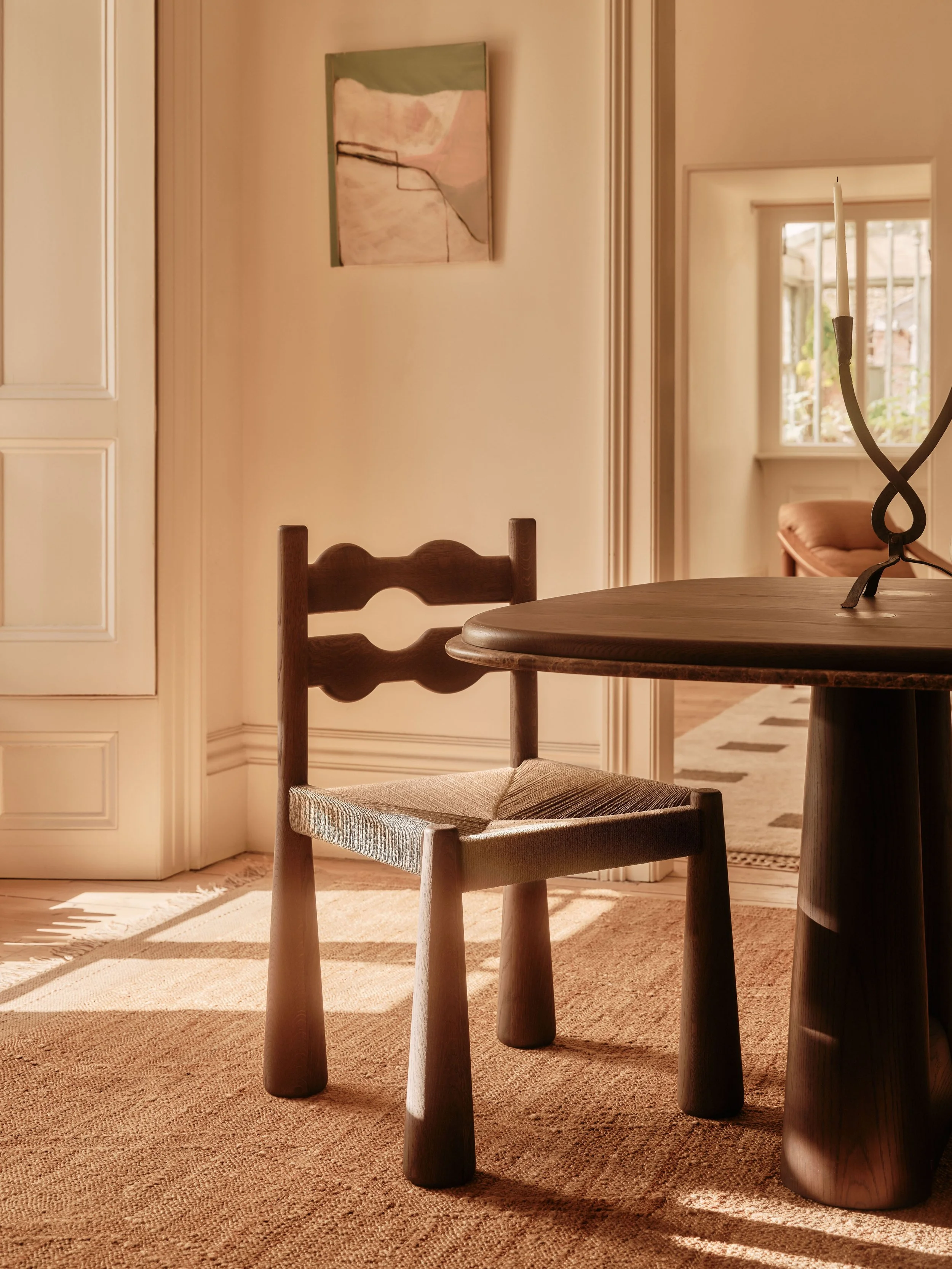 A wooden chair with a woven seat sits in front of a round wooden table. In the background, a beige wall with a small abstract painting, a window with white framing, and a partial view of a room with a built-in bench and a window are visible. The room