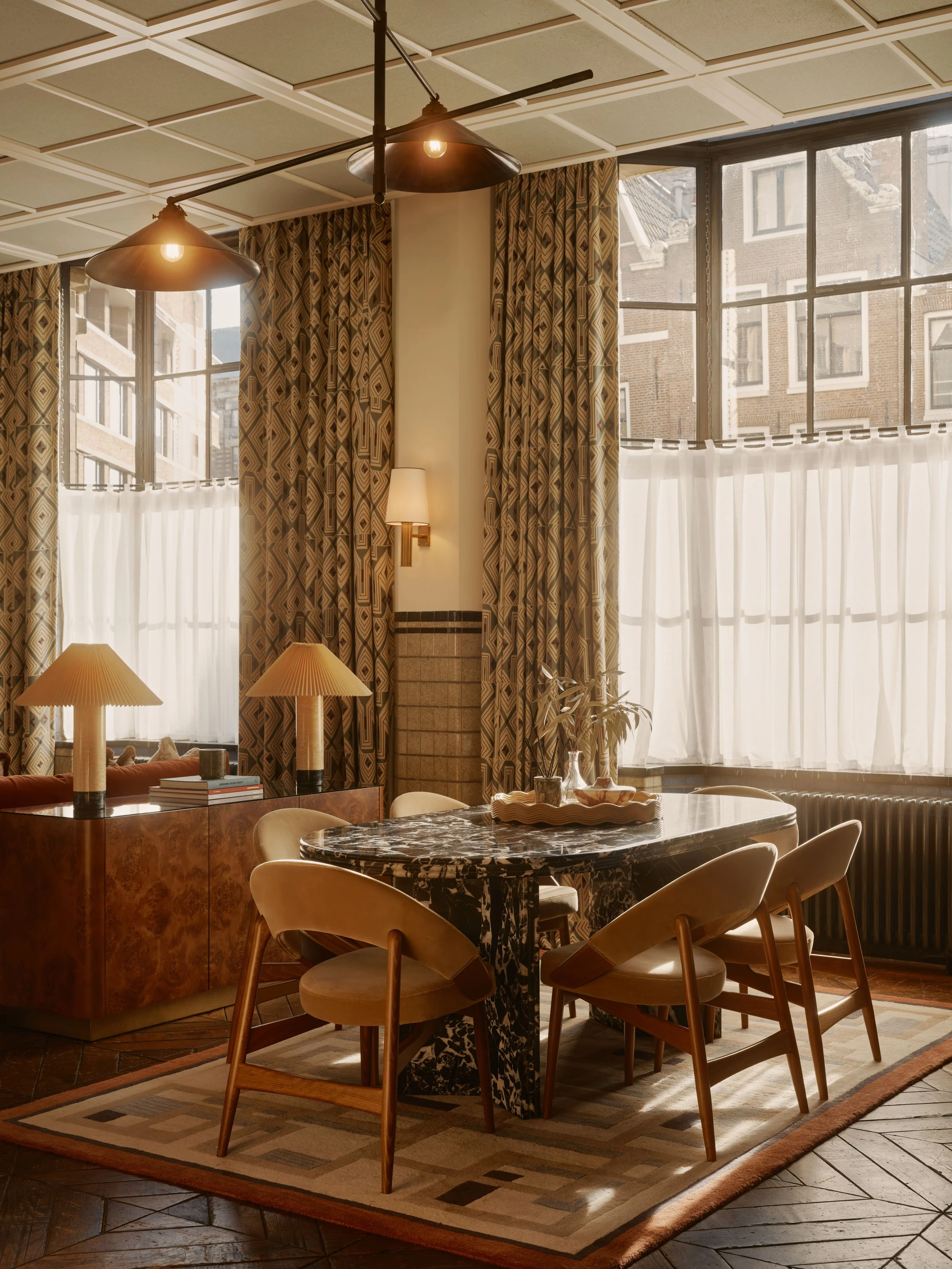 Interior of a sunlit dining room with a round marble table, six chairs, decorative curtains, a sideboard with lamps, and large windows showing neighboring brick buildings.