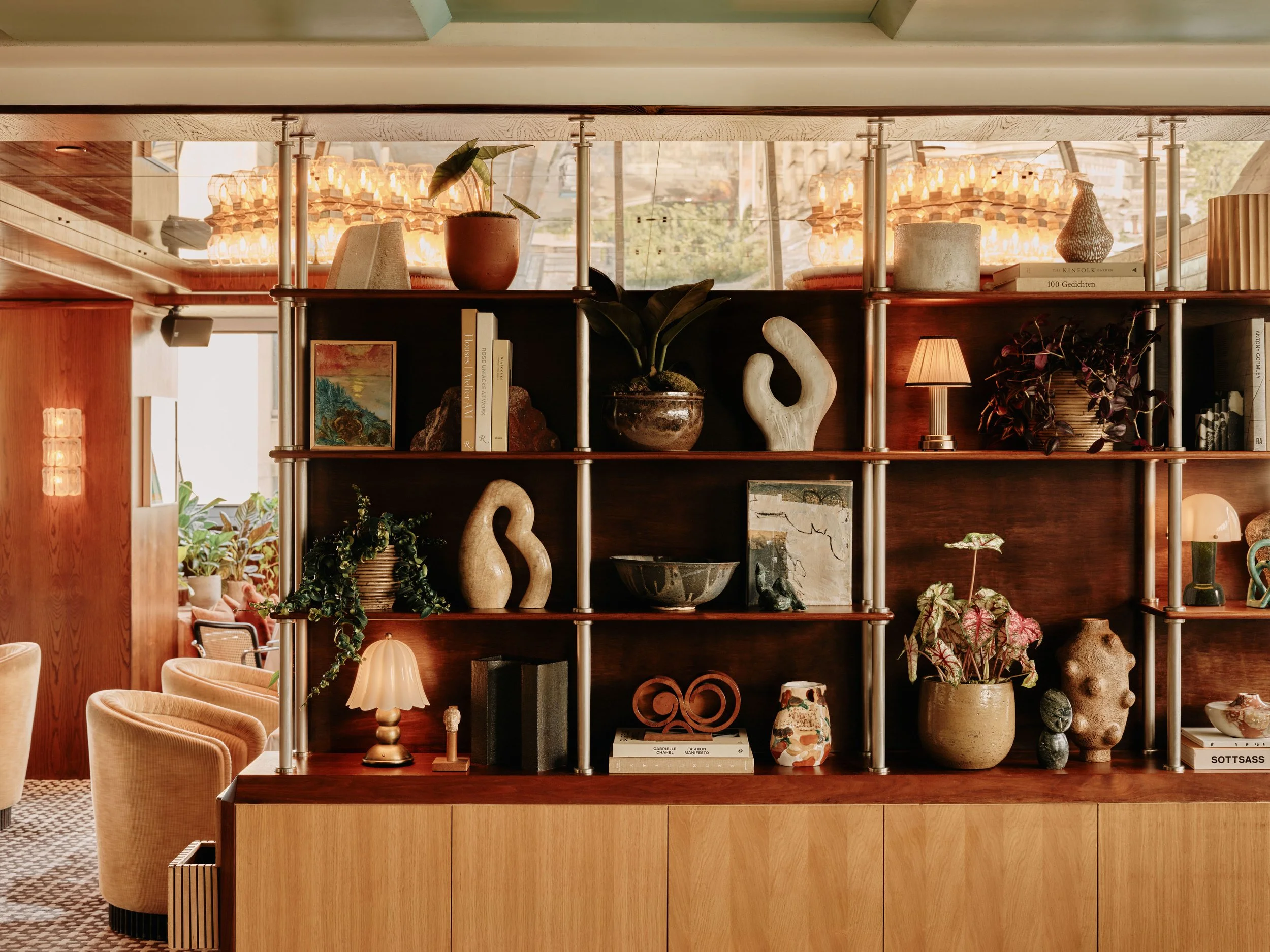Interior of a modern living space with a wooden shelving unit filled with decorative items, books, and plants, with warm lighting and large windows in the background. Captured at Soho House 180