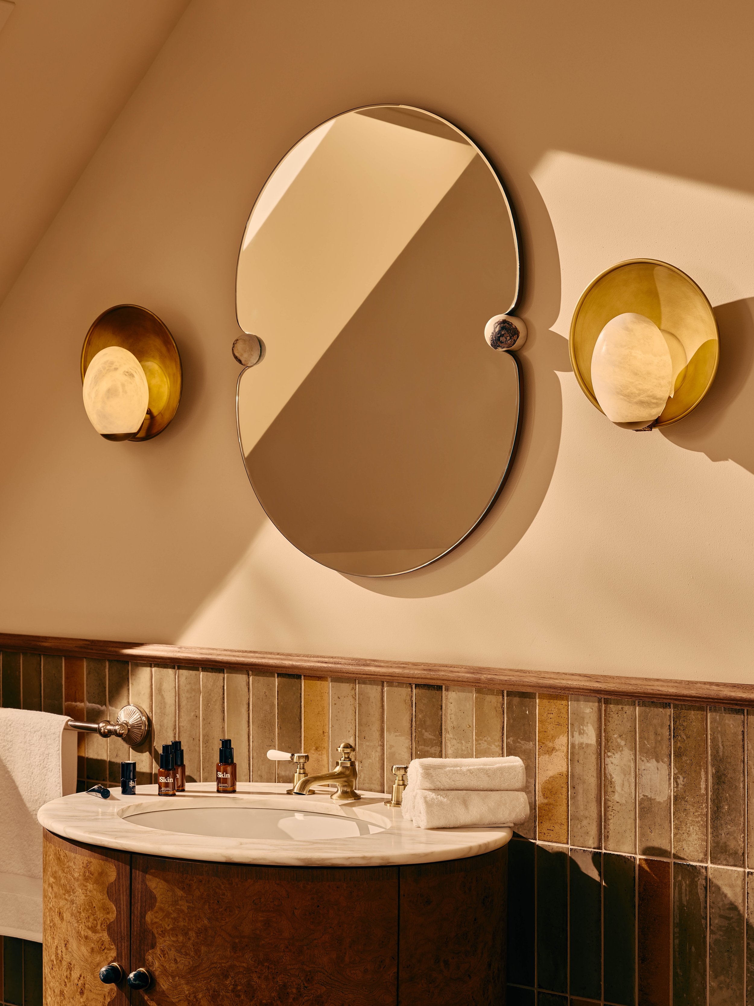 Bathroom vanity with a white marble countertop, gold faucet, and a round mirror on the beige wall. Two gold bowls with moon-shaped lamps and rocks, a stack of white towels, and bottles of skincare products are on the counter. There are beige and brow