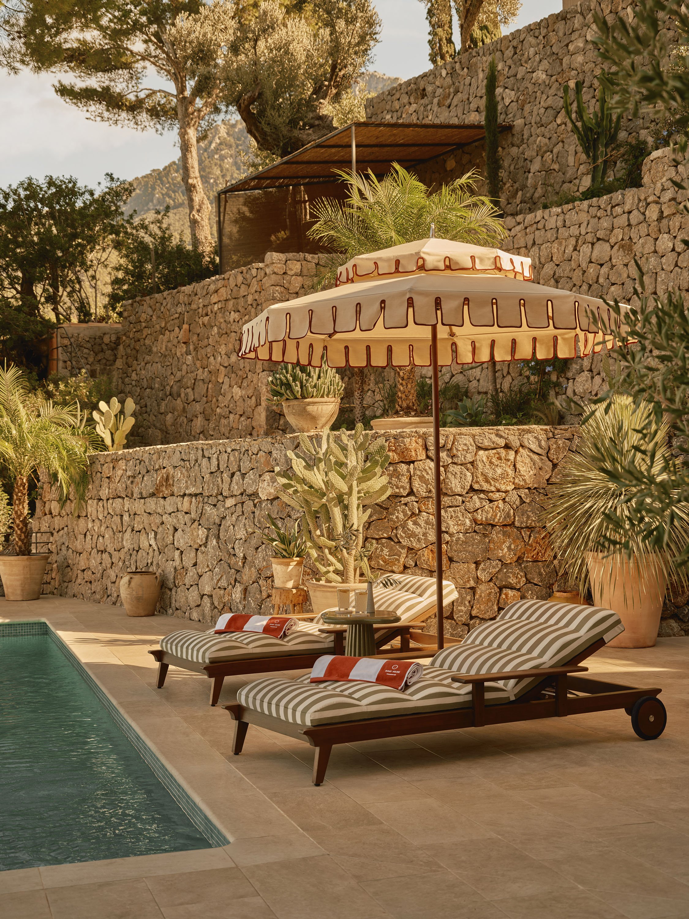 A backyard with a swimming pool, two striped lounge chairs with towels, a large umbrella, a stone wall, and various potted plants under trees, with a mountain in the background.